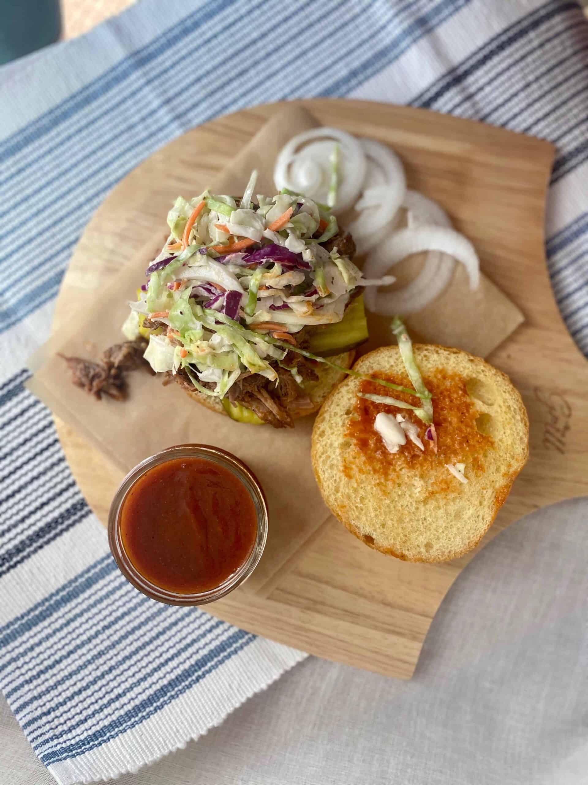 Overhead view of a pulled pork sandwich topped with colorful coleslaw and pickles on a wooden board, with a side of sliced onions and a small glass of barbecue sauce nearby.