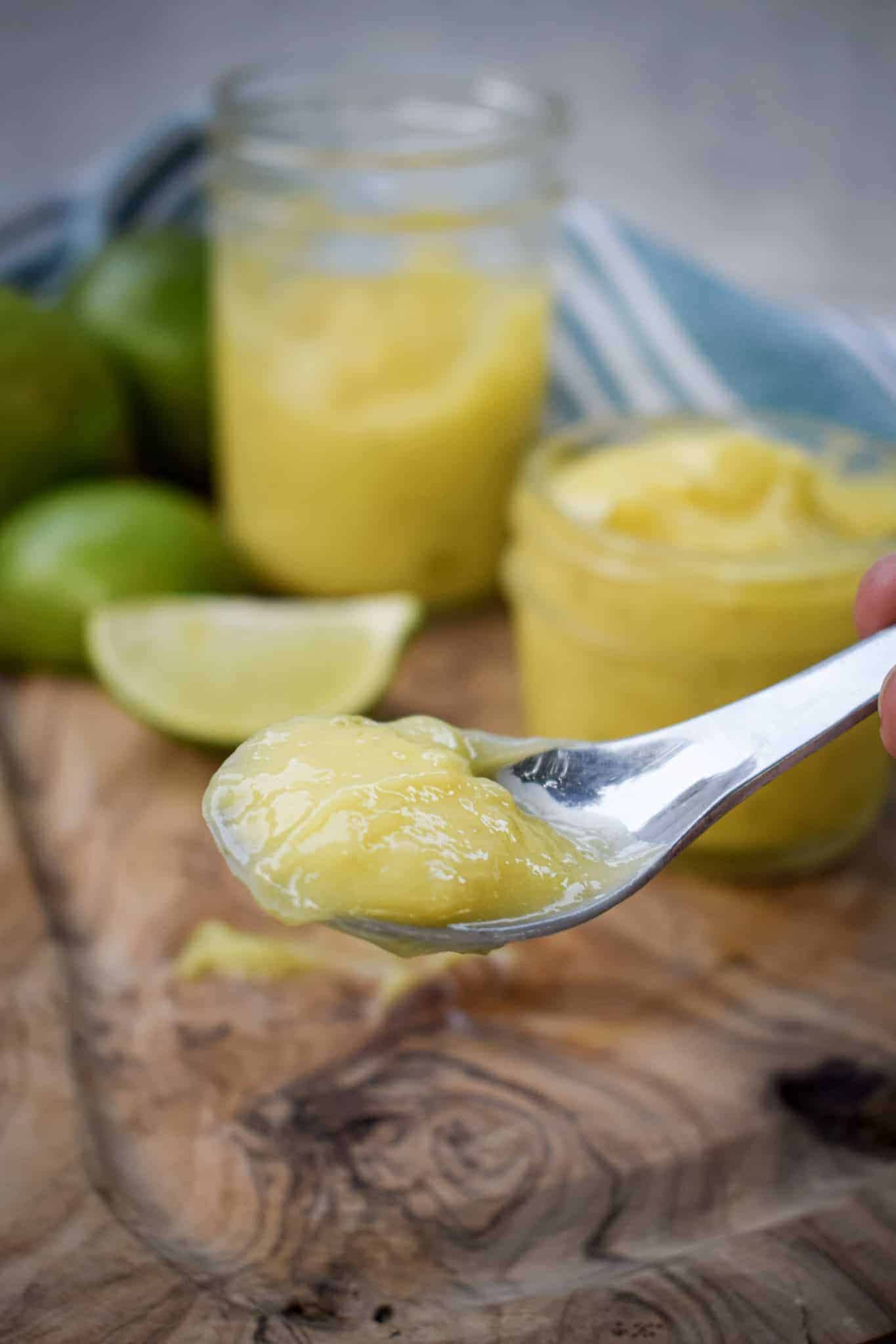 Close-up of a spoonful of creamy homemade lime curd with jars of lime curd and fresh limes in the background.