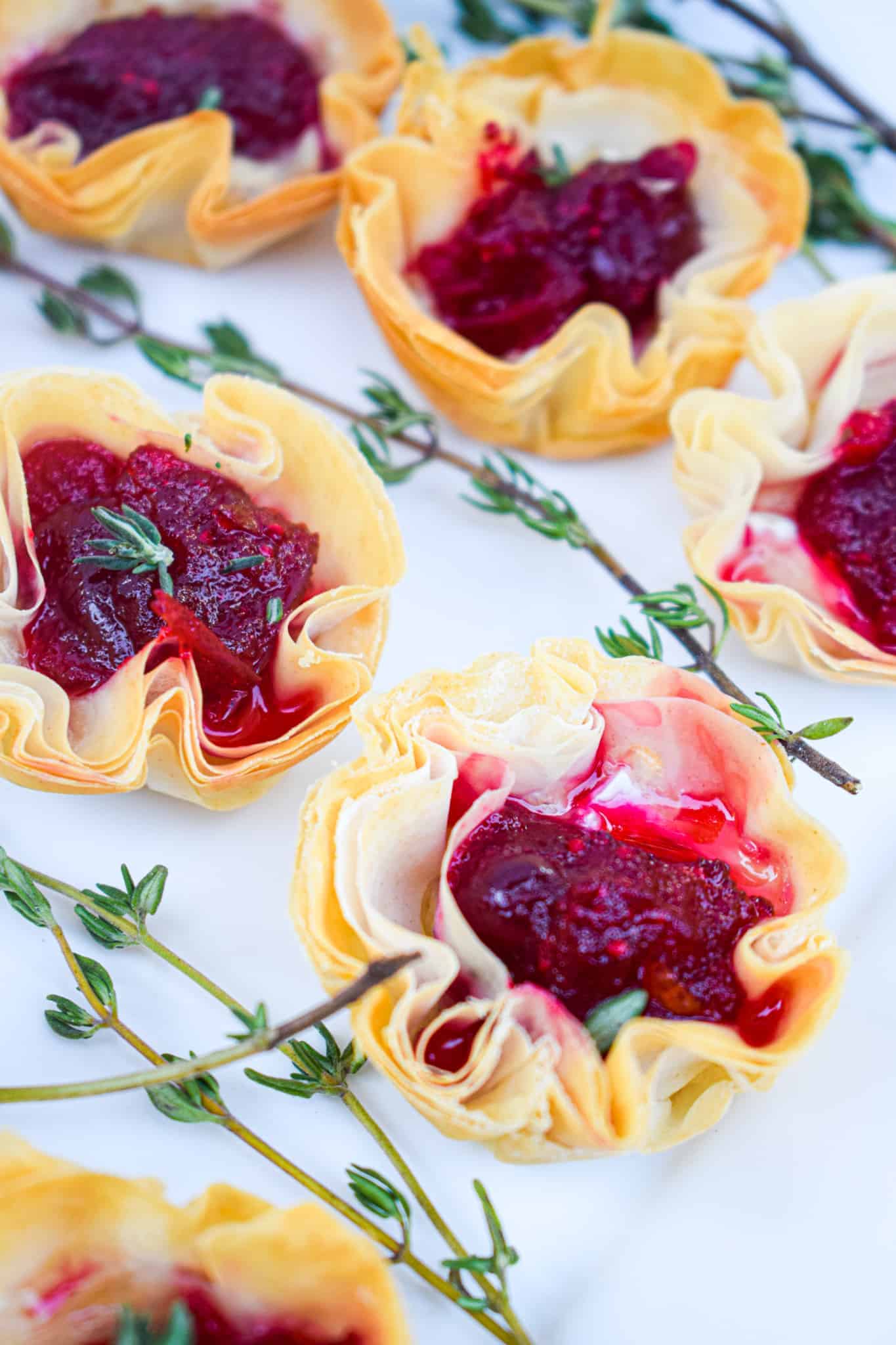 Mini Bri & Cranberry Phyllo Tarts filled with red cranberry sauce and garnished with small thyme sprigs, arranged beautifully on a white surface.