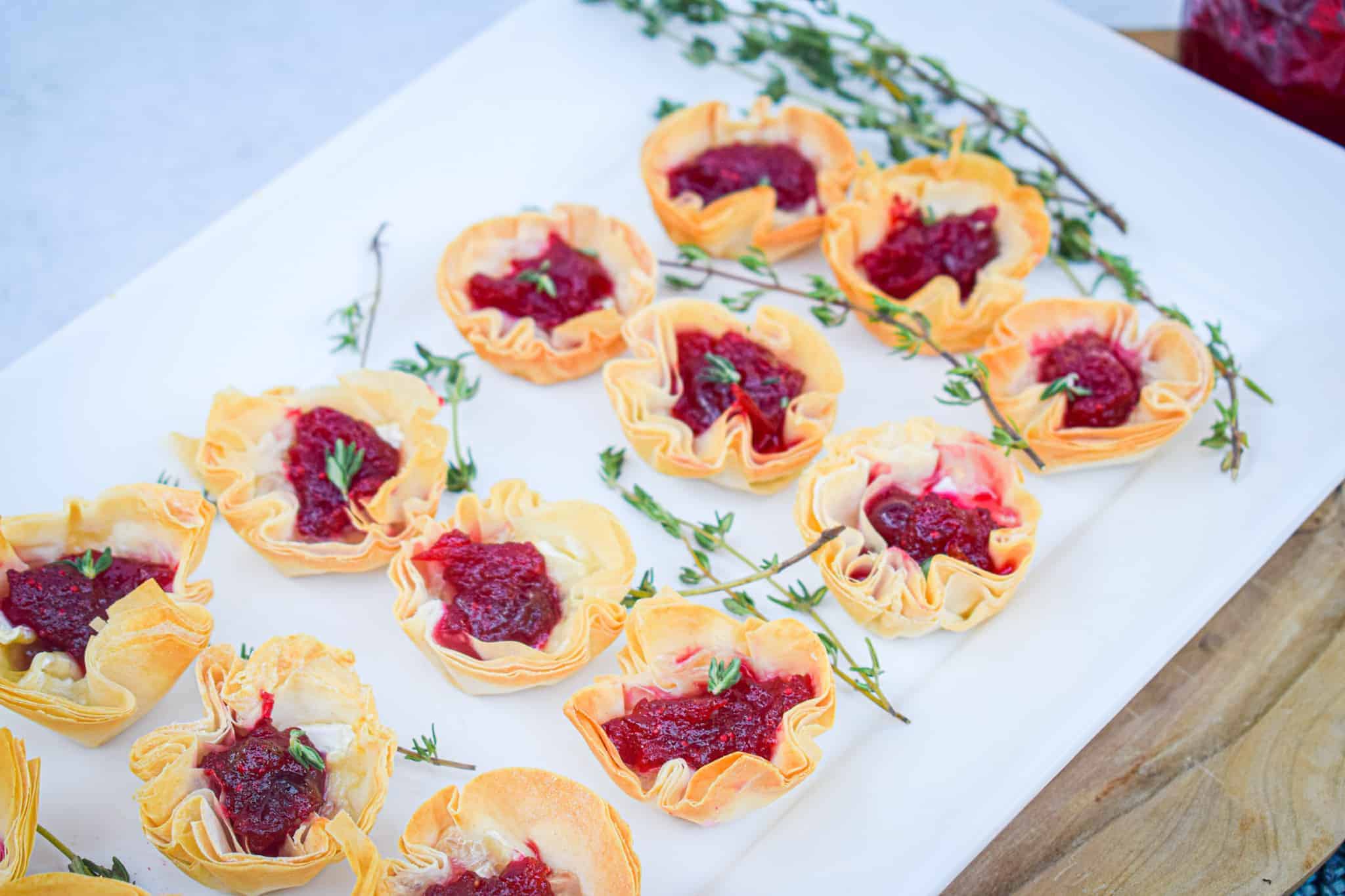 Mini Bri & Cranberry Phyllo Tarts filled with red cranberry sauce and garnished with small green herbs, arranged on a white tray with sprigs of fresh thyme.