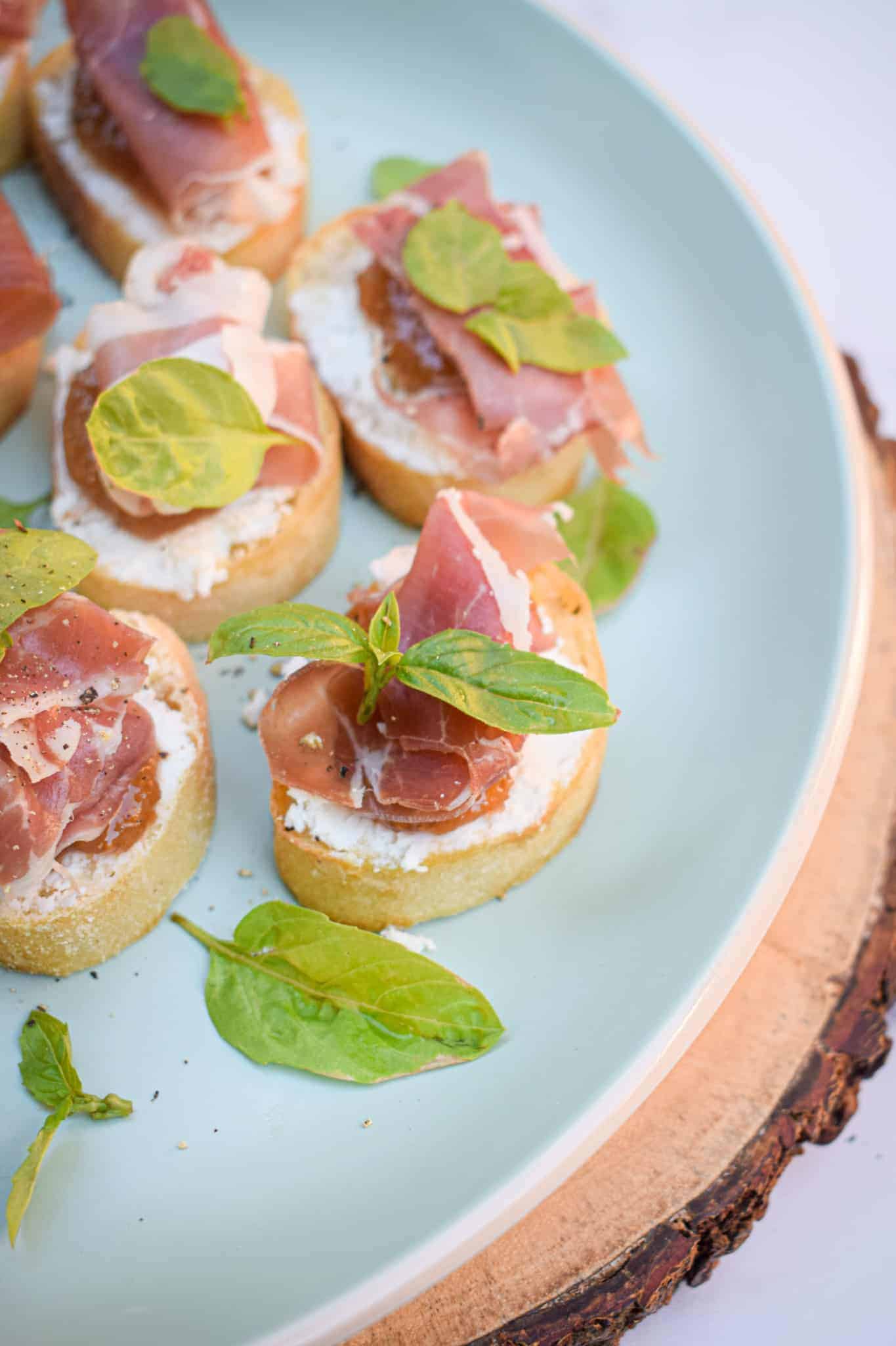 A close of up a plate of crostini topped with goat cheese, fig jam, olive oil and fresh basil. 