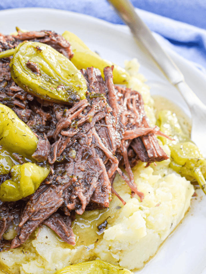 Whole30 Mississippi pot roast—shredded and topped with pepperoncini peppers—served over a bed of mashed potatoes on a white plate with a fork.