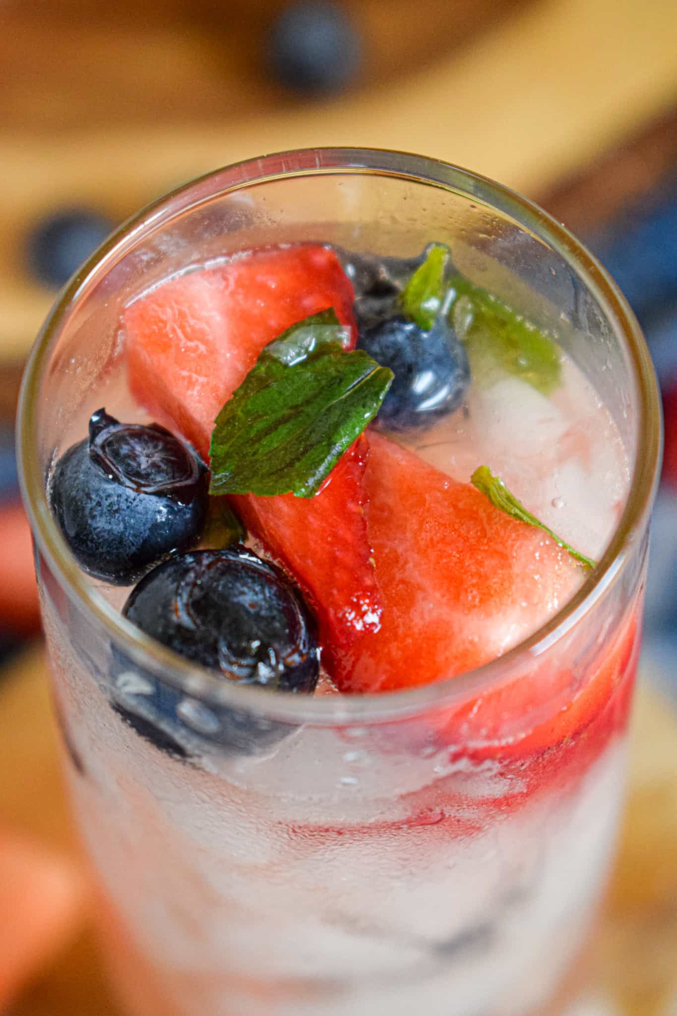 A close-up of a 4th of July mocktail, featuring a glass filled with ice, blueberries, strawberry slices, and mint leaves.