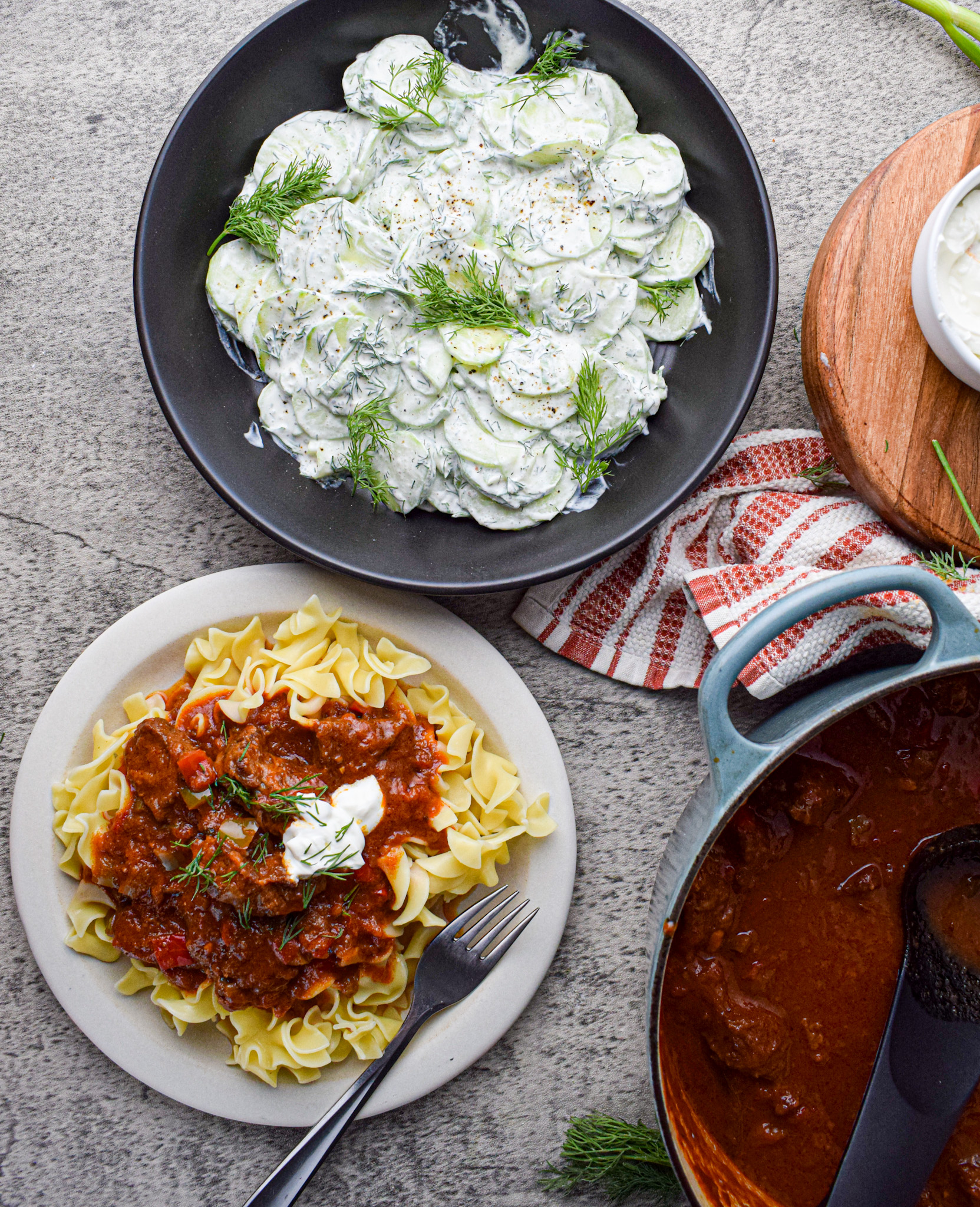 Overhead view of creamy Polish cucumber salad (Mizeria) garnished with dill in a black bowl, next to a plate of beef goulash over egg noodles with sour cream and a pot of tomato-based stew.