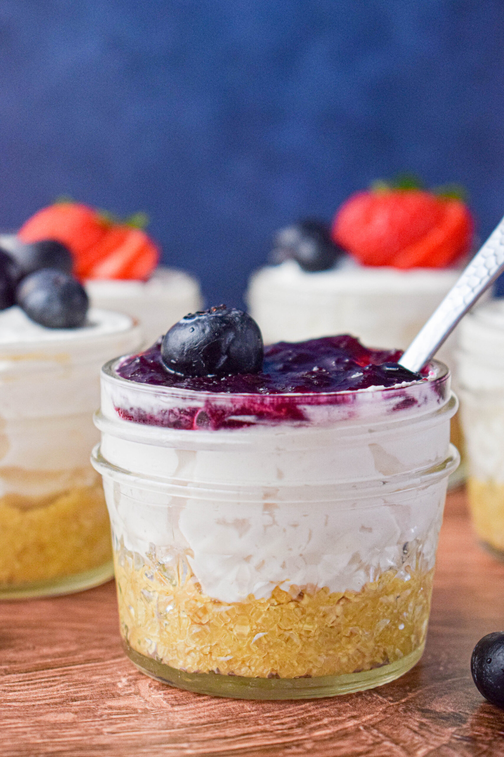 individual serving of cheese cake in a mason jar topped with jam and a blueberry with a spoon in the jar 