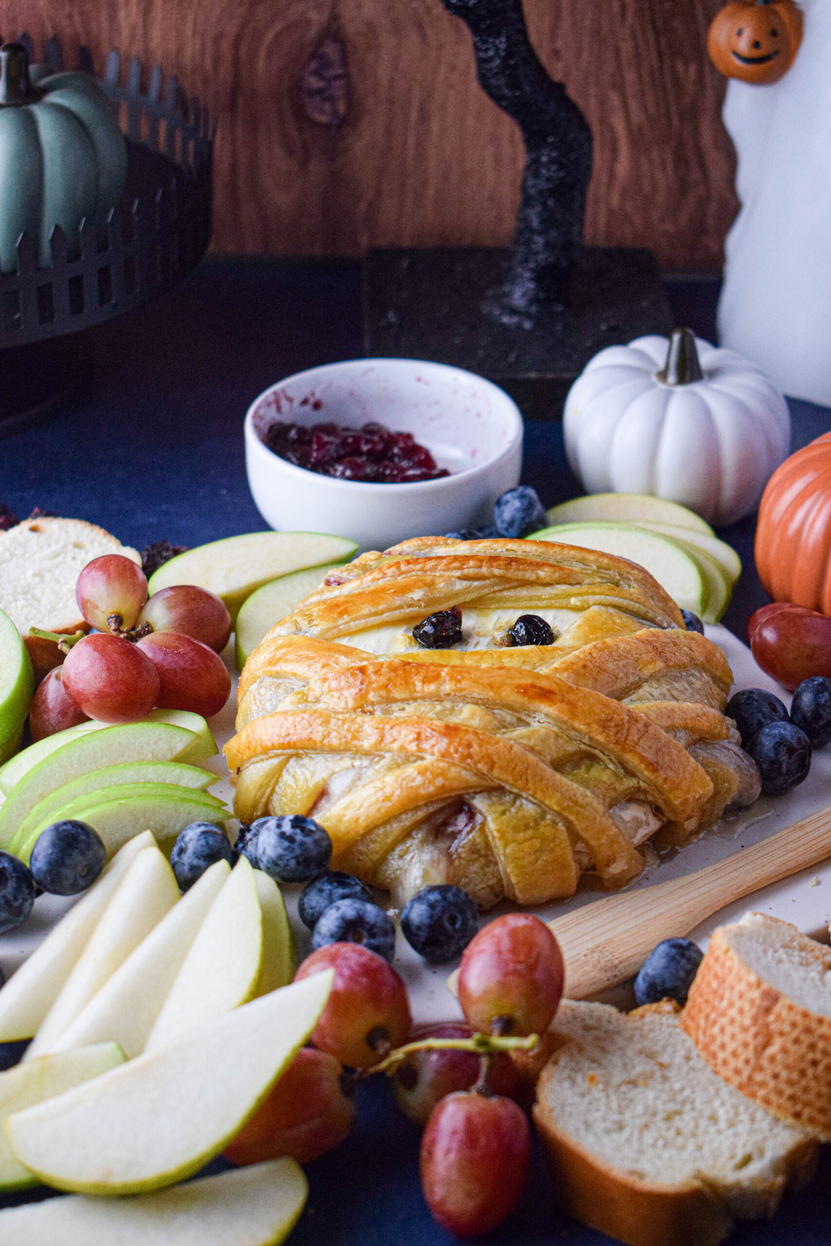 spooky halloween background with a mummy puff pastry wrapped baked brie surrounded by fruit and bread