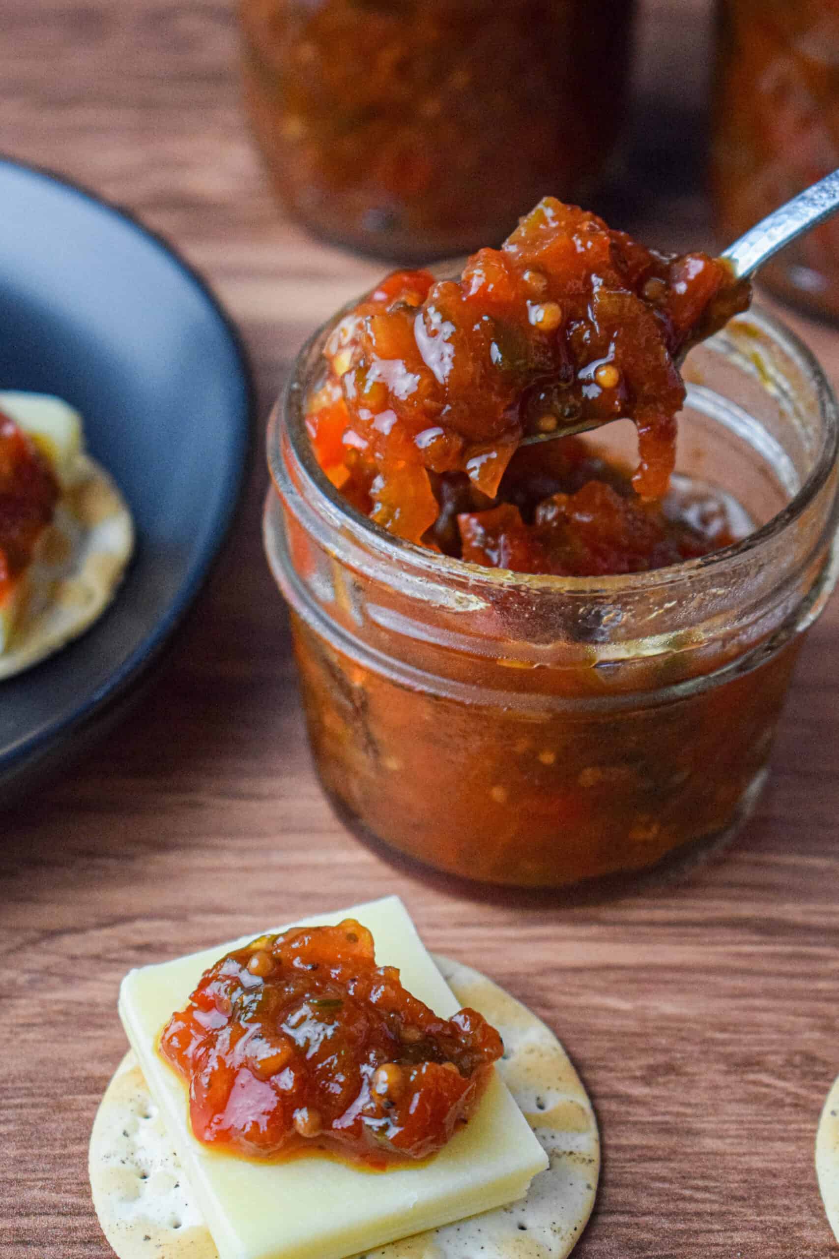 mason jar full of tomato chutney next to a cracker with cheese topped with chutney 