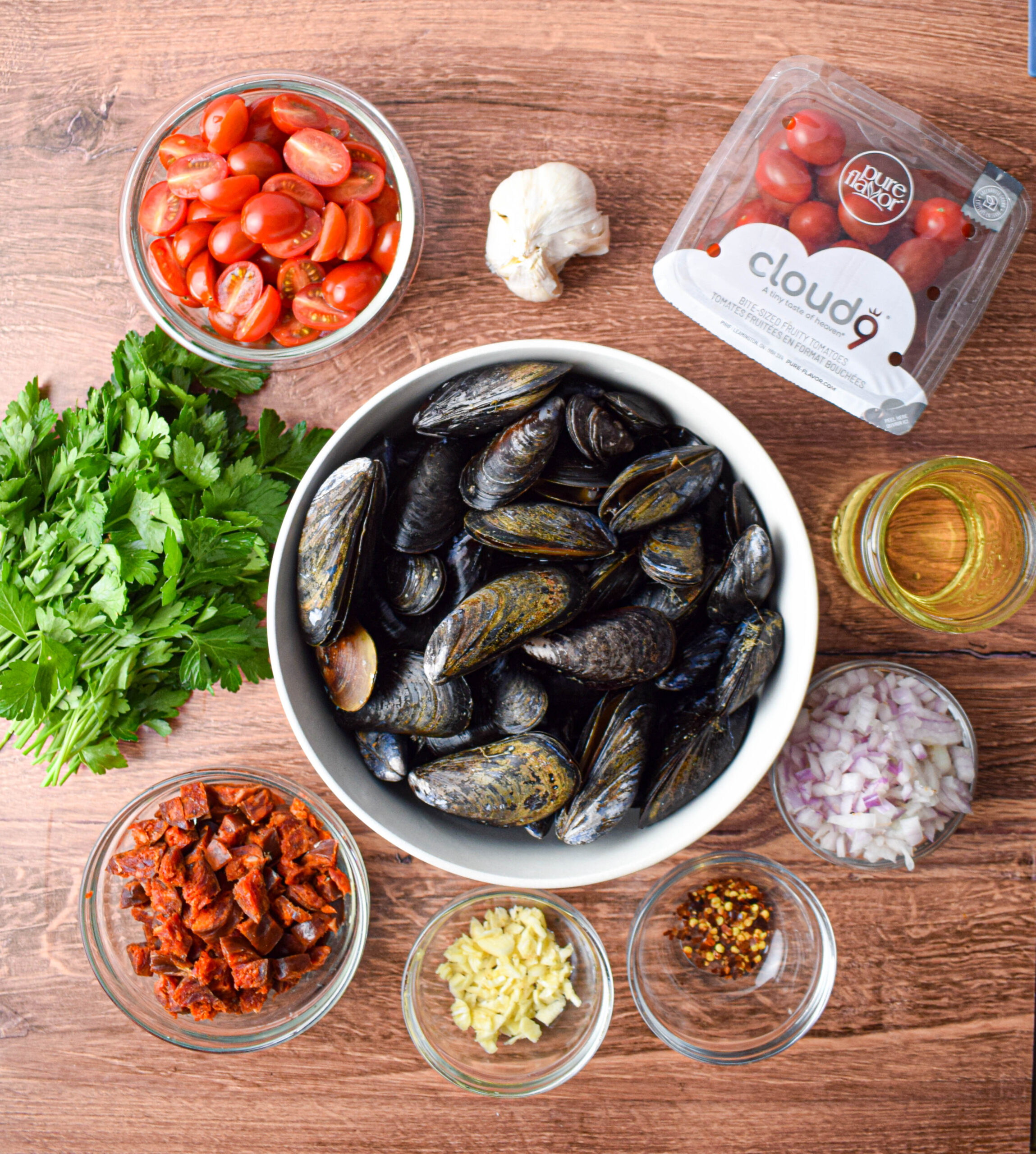 ingredients for chorizo and tomato steamed mussels. Bowl of halved tomatoes, garlic, fresh parsley, bowl of diced Spanish chorizo, bowl of diced shallot, glass of white wine, large bowl full of fresh mussels