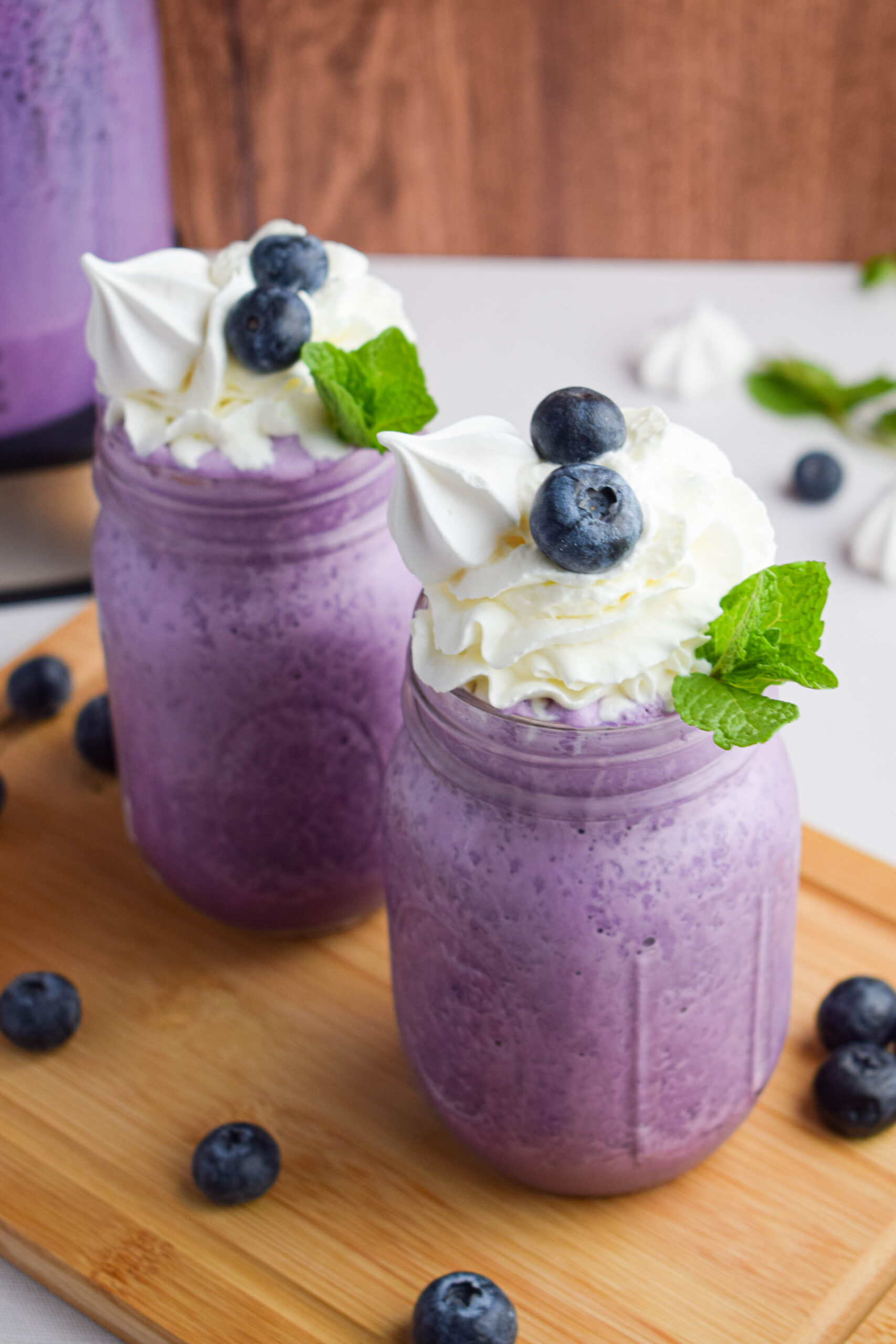two blueberry milkshakes in a mason jars topped with whipped cream, a meringue cookie, fresh blueberries and a sprig of mint 