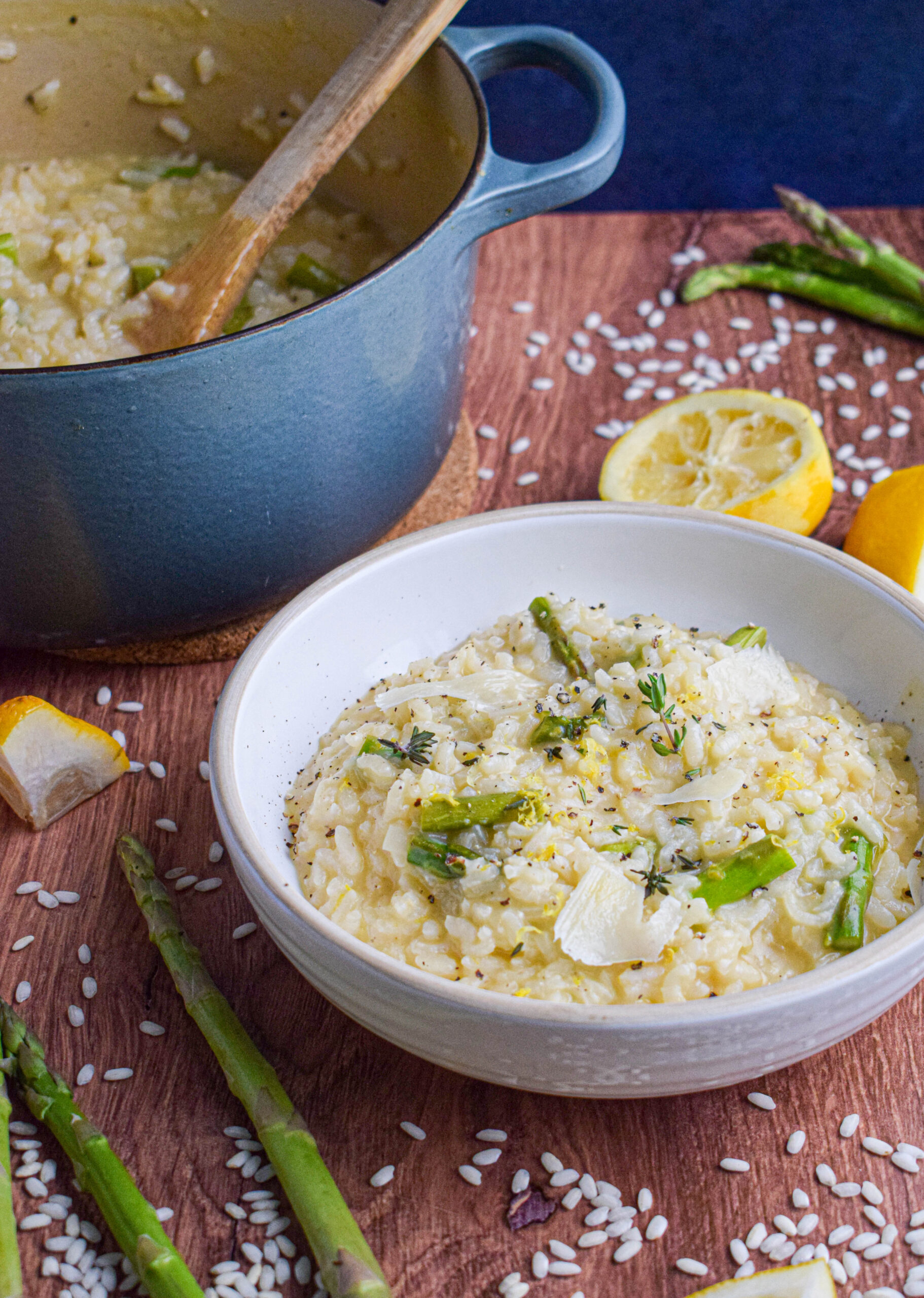 white bowl of spring lemon parmesan risotto surrounded by lemons and asparagus next to dutch oven full of risotto