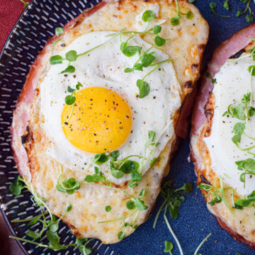 A Croque Madame featuring two slices of toasted bread with melted cheese, ham, a sunny-side-up egg, and fresh microgreens on a dark blue plate.
