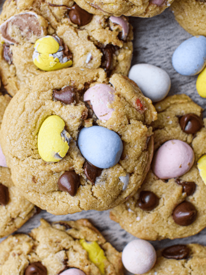 Mini Egg Cookies: Chocolate chip cookies topped with pastel candy-coated chocolate eggs, arranged on a wooden surface.