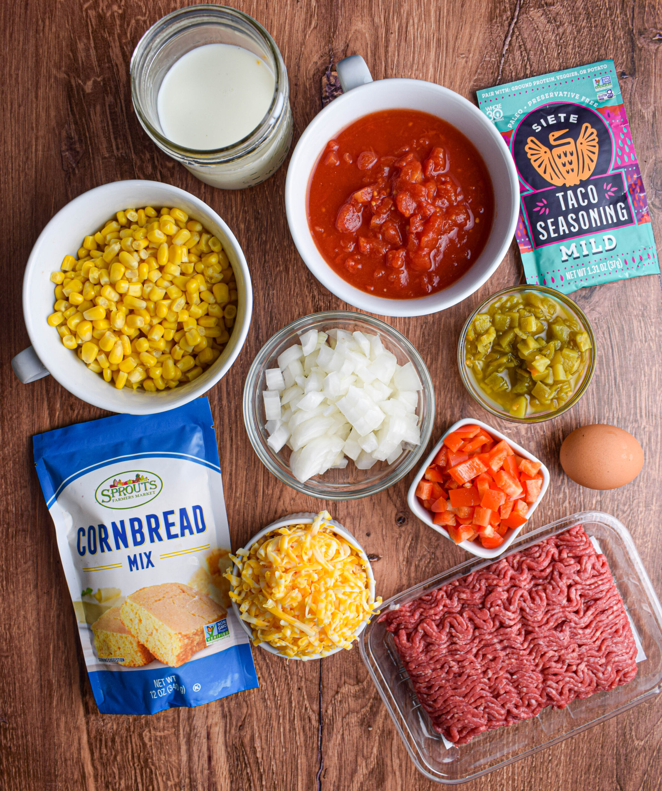 ingredients for beef tamale pie laid out in bowls on a wood surface, cornbread mix, canned corn, diced onions, green chiles, red peppers, taco seasoning, ground beef, shredded cheese, egg, milk