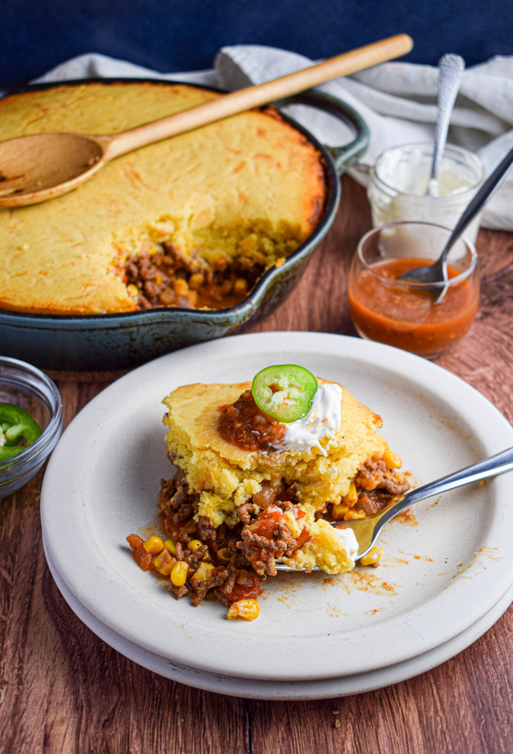 cast iron skillet of cooked tamale casserole and in front is a plated serving of the tamale pie with sour cream, salsa and a sliced jalapeno 