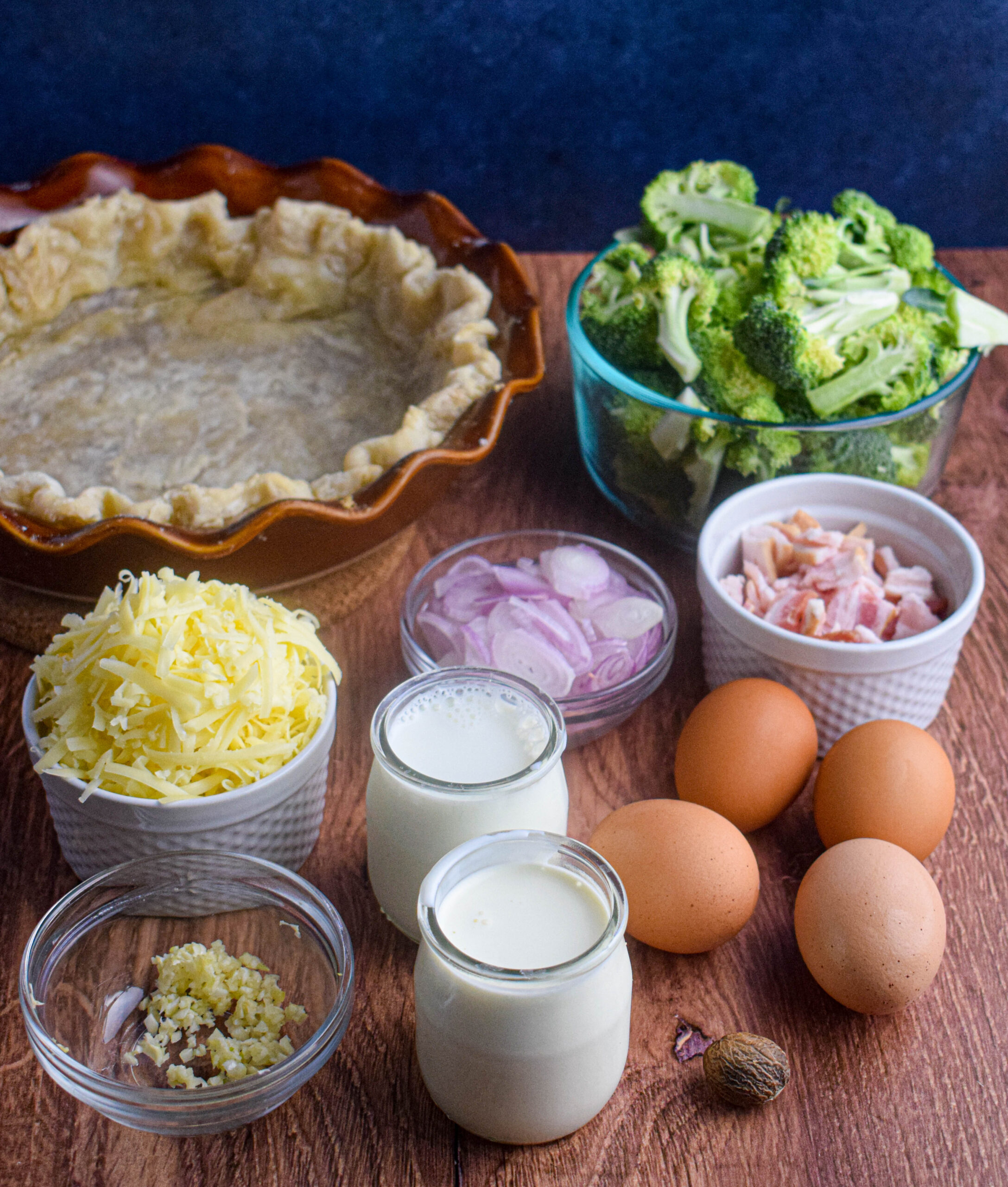 ingredients for broccoli cheddar quiche: eggs, heavy cream, milk, garlic, cheese, shallot, bacon, broccoli and pie crust 