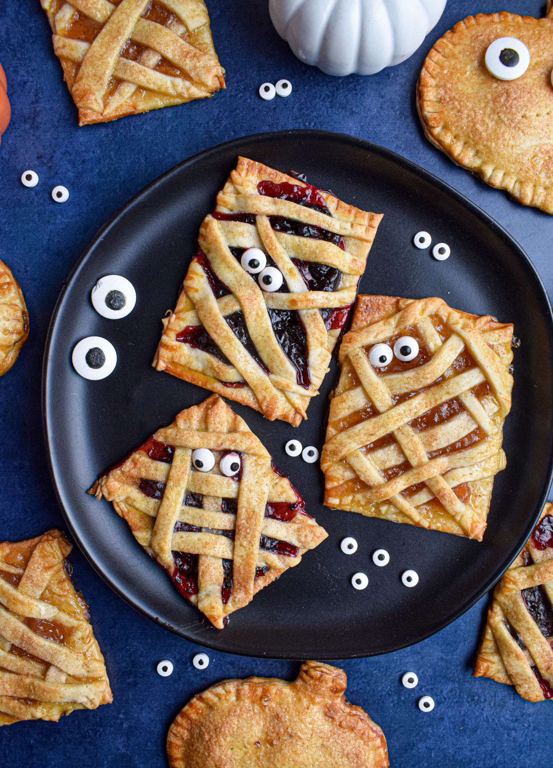 black plate on a blue surface with mummy hand pies surrounded by candy eyes and pumpkin Halloween hand pies