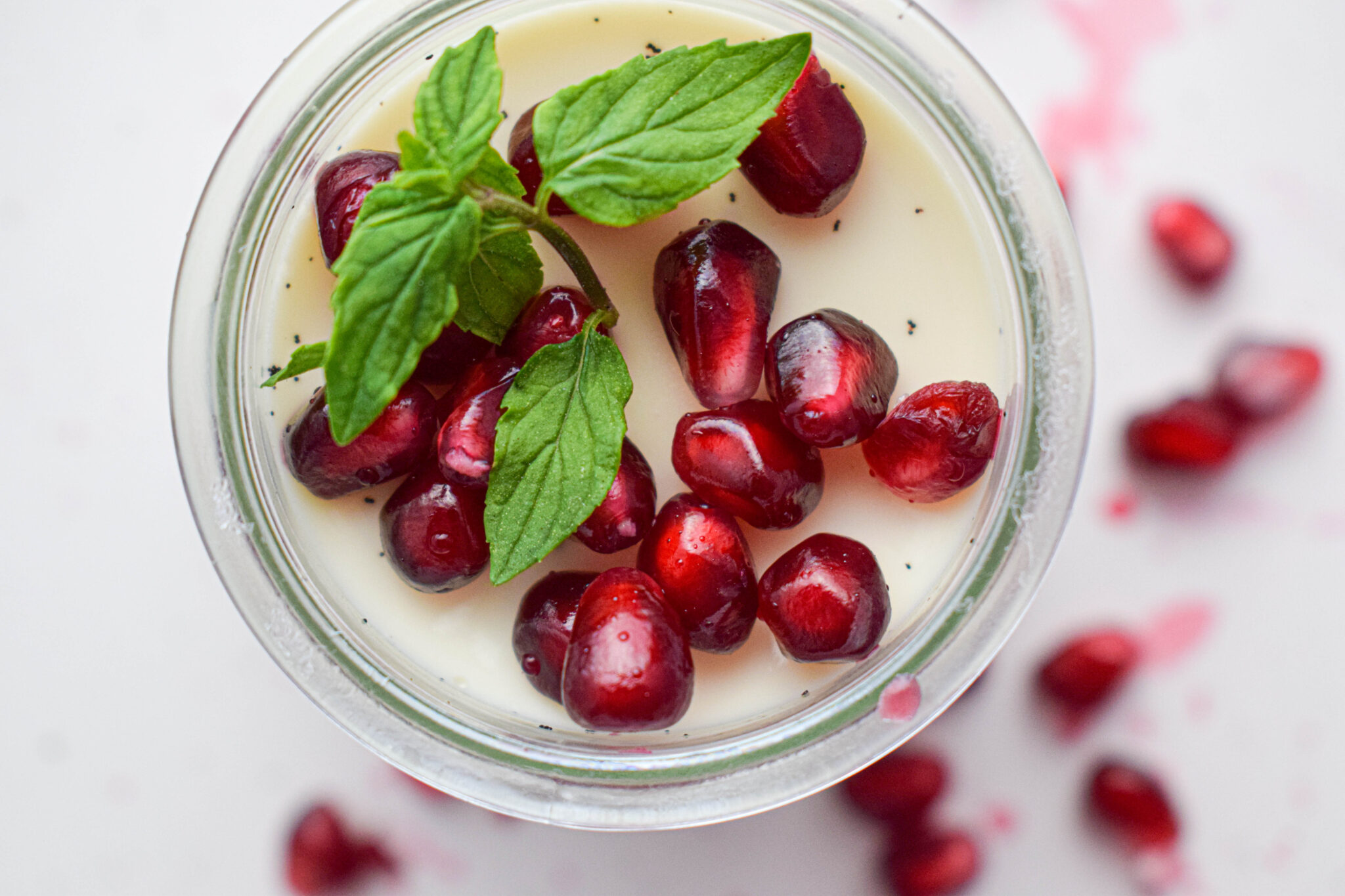 A glass of creamy dessert topped with pomegranate seeds and fresh mint leaves. More pomegranate seeds are scattered around the glass on a light surface.