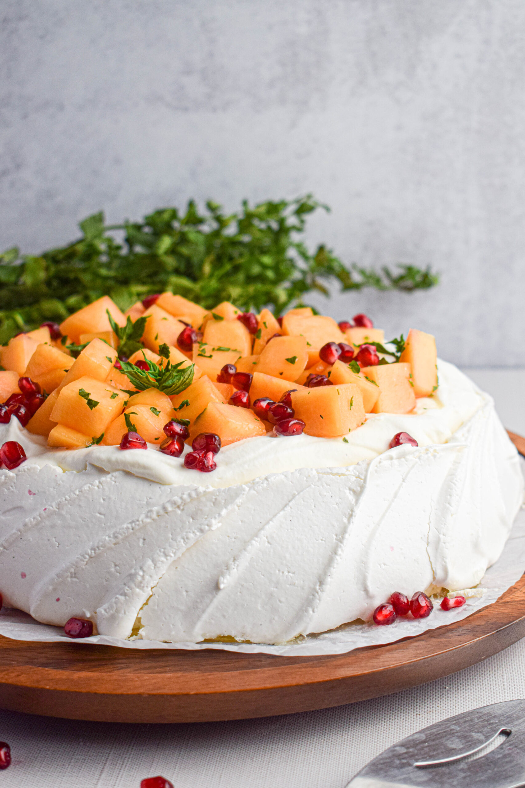 side shot of a pavlova topped with whipped cream, canteloupe and pomegranate seeds with fresh mint