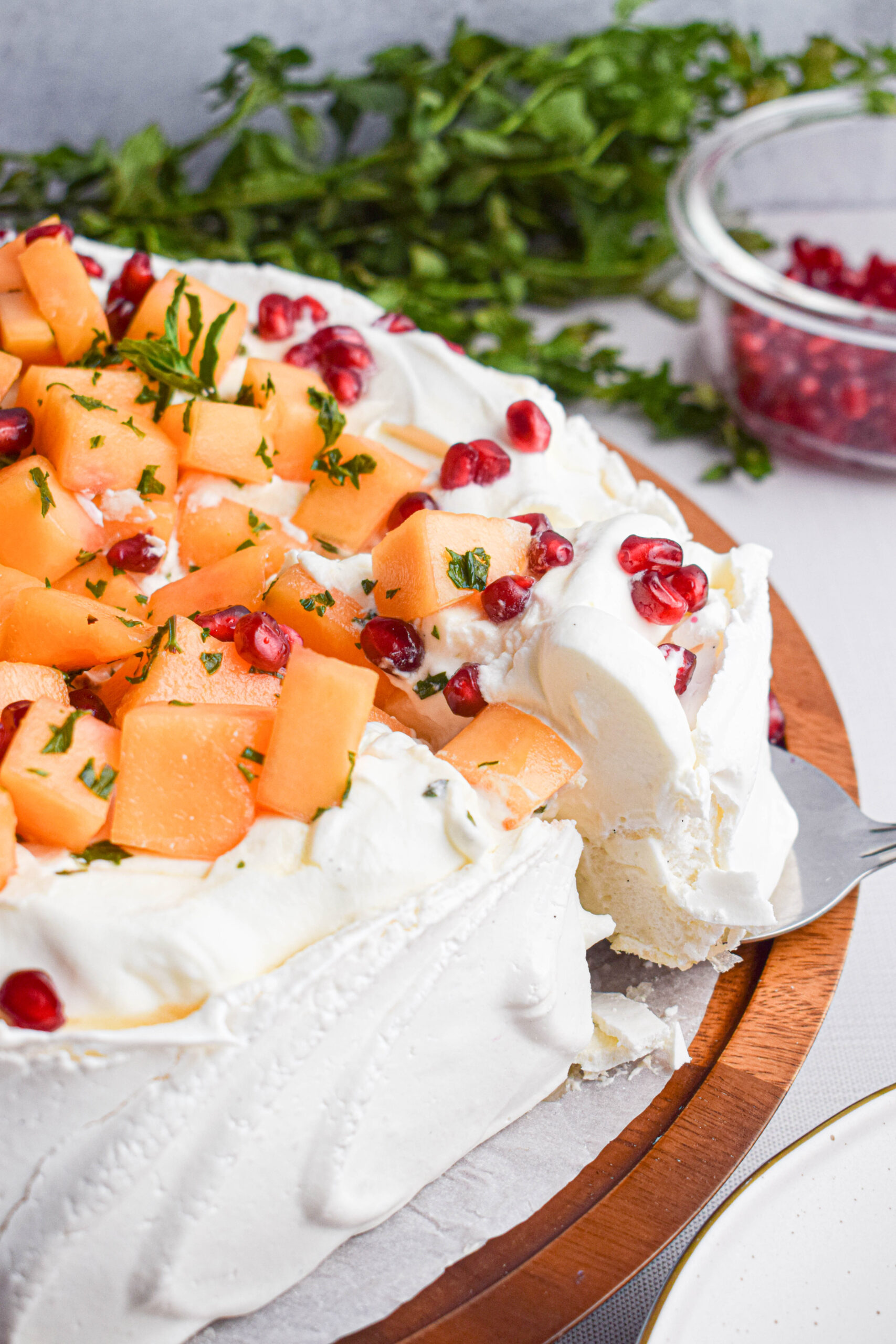 side shot of a melon pavlova topped with cantaloupe and pomegranate with a serving spoon removing a piece