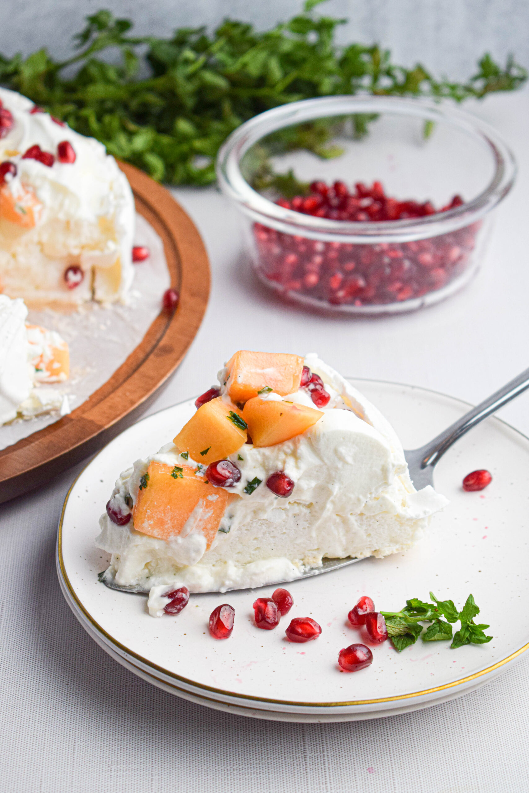 slice of melon pavlova on a plate with whipped cream, cantaloupe melon and pomegranate seeds, garnished with fresh mint. Behind there is a bowl of pomegranate seeds 