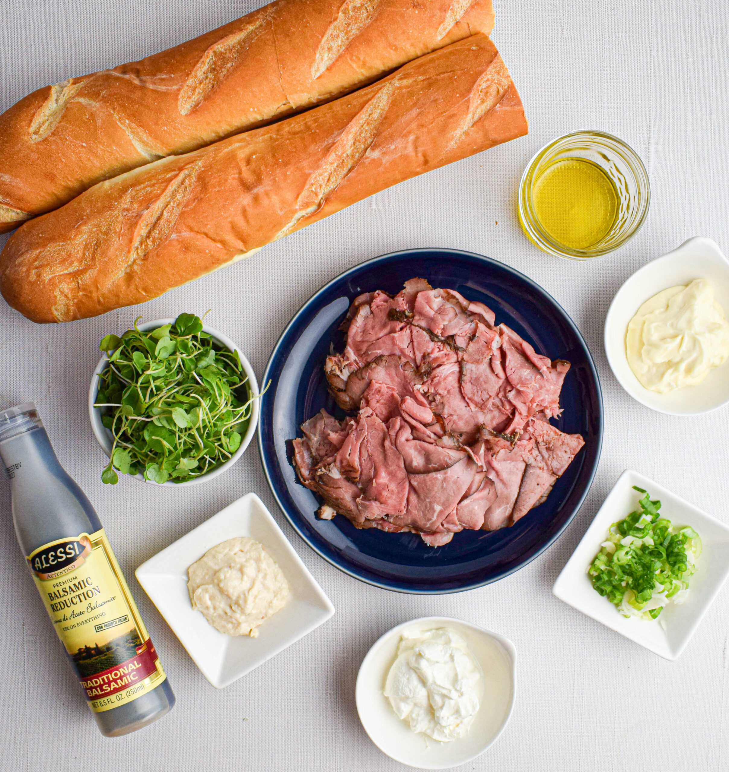 ingredients laid out for roast beef crostini on plates and in bowls: shaved roast beef, micro arugula, horseradish, sour cream, mayonnaise, sliced green onions, balsamic reduction, olive oil, baguette
