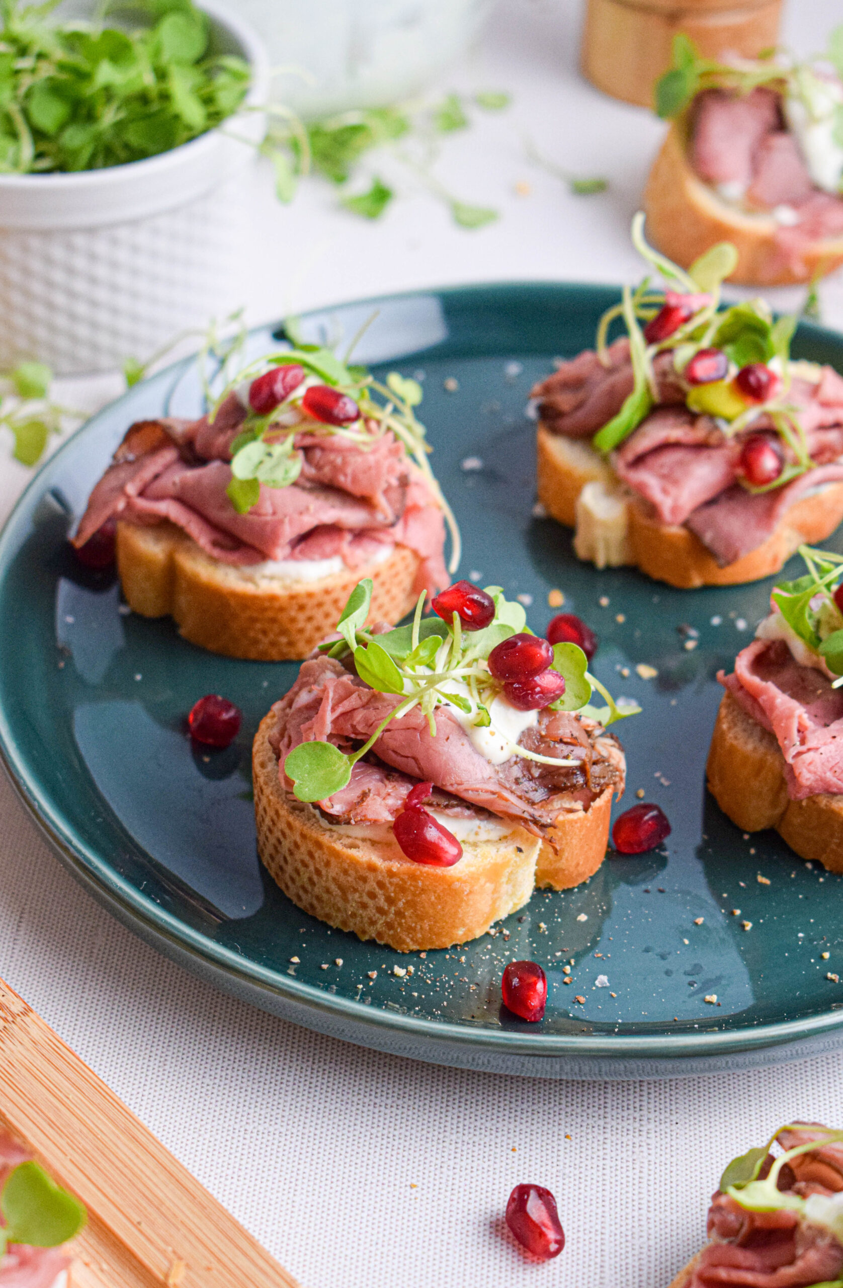 green plate with crostini topped with roast beef, horseradish sauce, micro arugula and pomegranate seeds 
