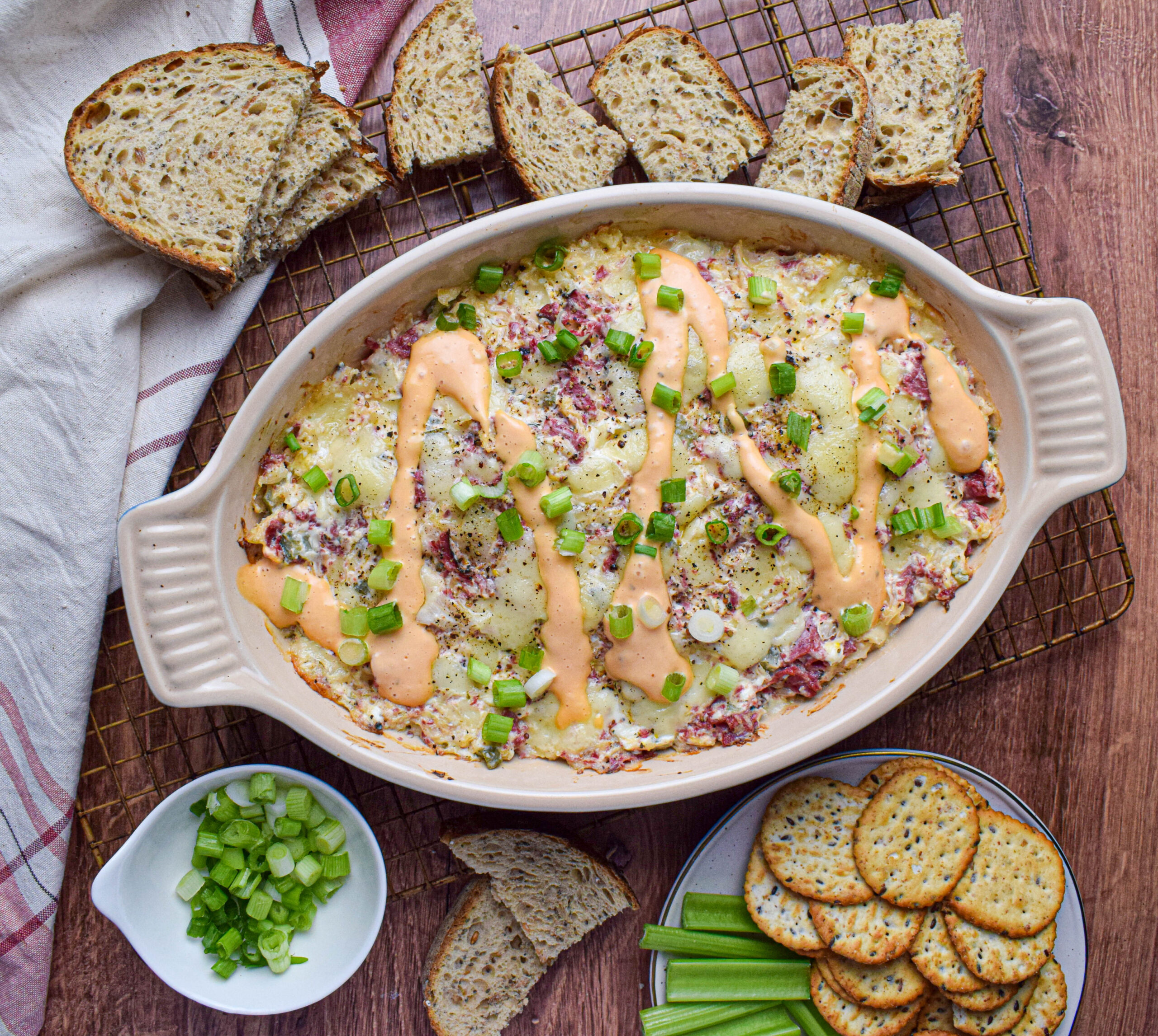 Baked reuben sandwich tailgating dip topped with thousand island dressing and green onions surrounding by toasted rye bread slices, crackers and celery.
