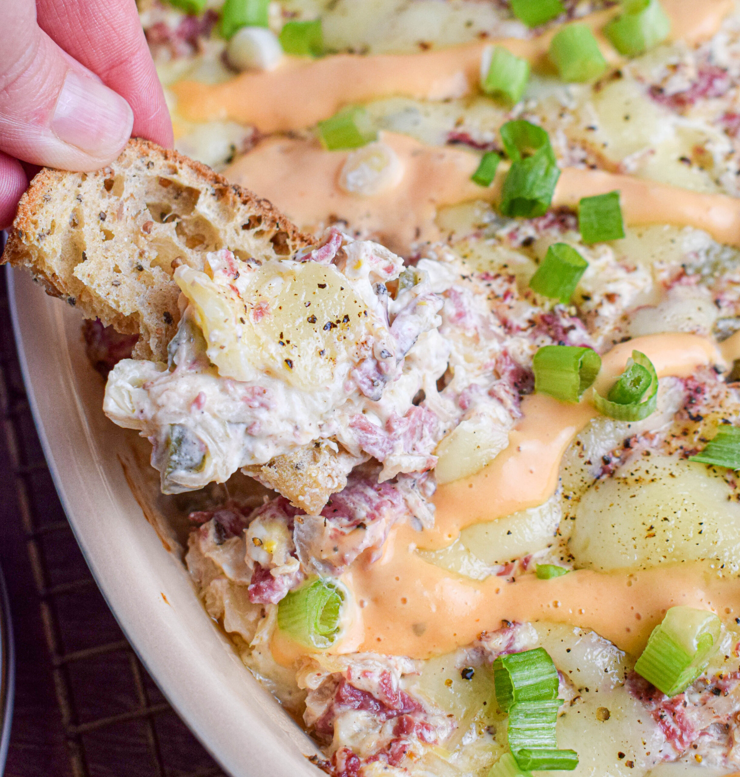 A piece of rye bread is being dipped into a dish of hot reuben dip with melted cheese, topped with thousand island dressing and sliced green onions.
