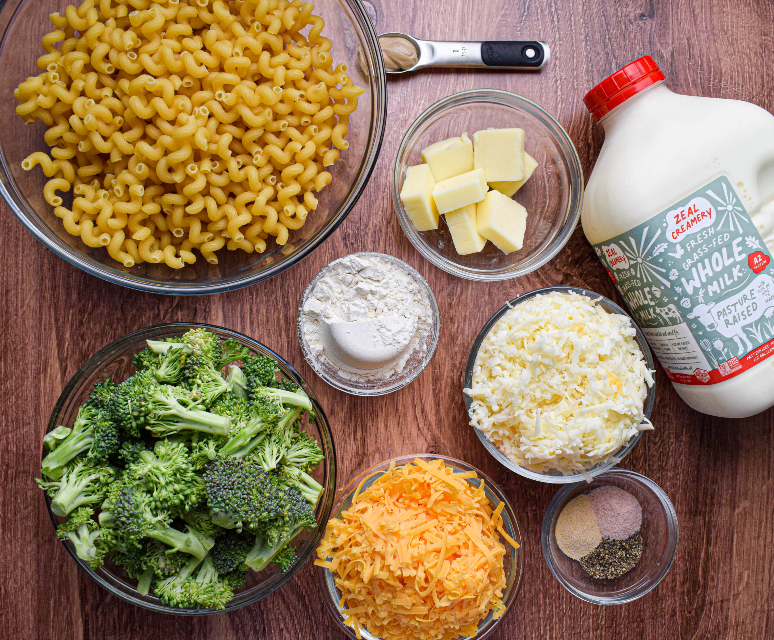 Ingredients for stovetop broccoli mac and cheese are laid out on a wood surface: cavatappi pasta, butter, mustard, milk, flour, two types of cheese, seasoning and fresh broccoli florets.