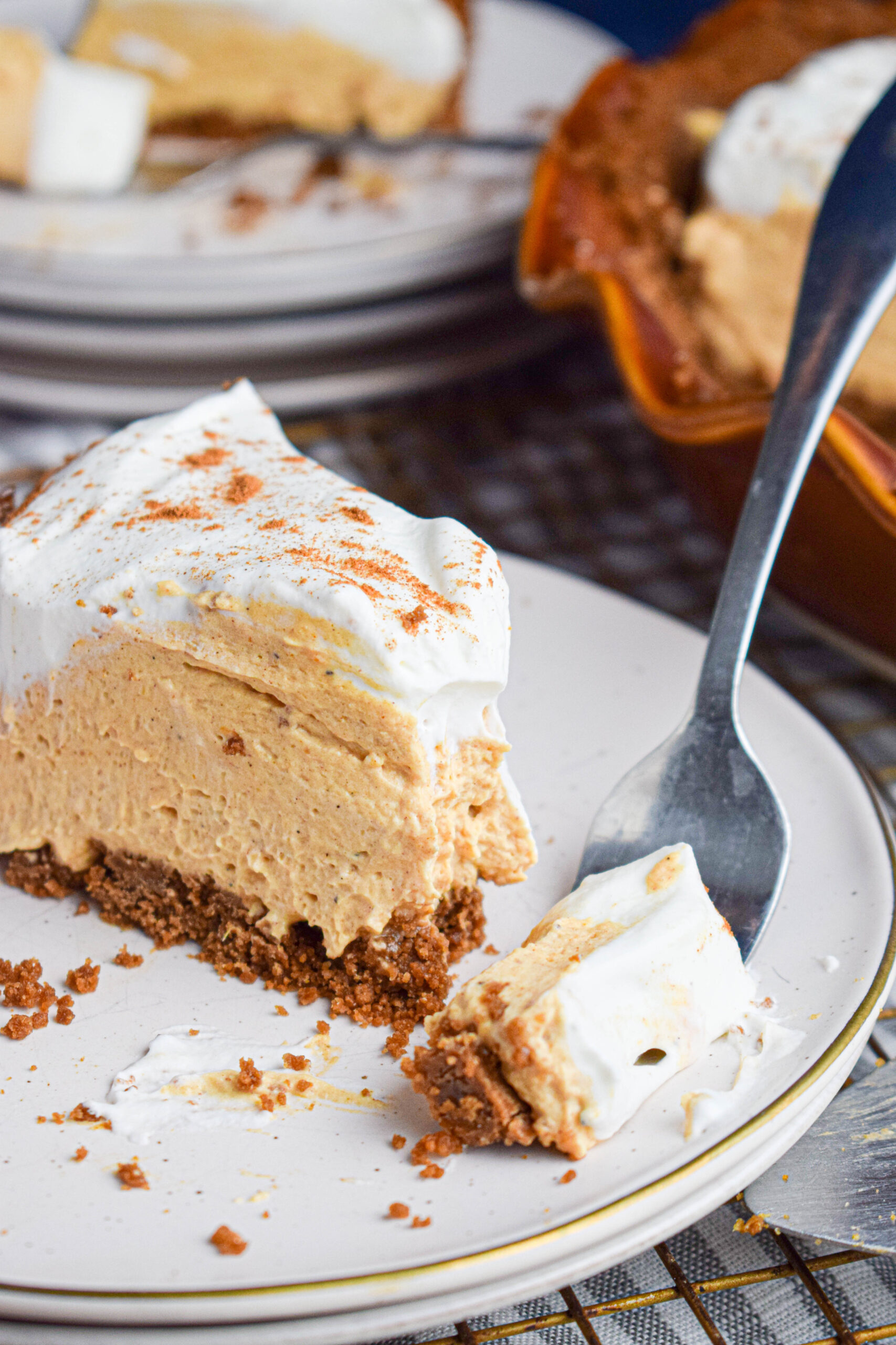 A slice of no bake pumpkin cheesecake pie on a white plate topped with whipped cream with a fork taking out a layered bite. 