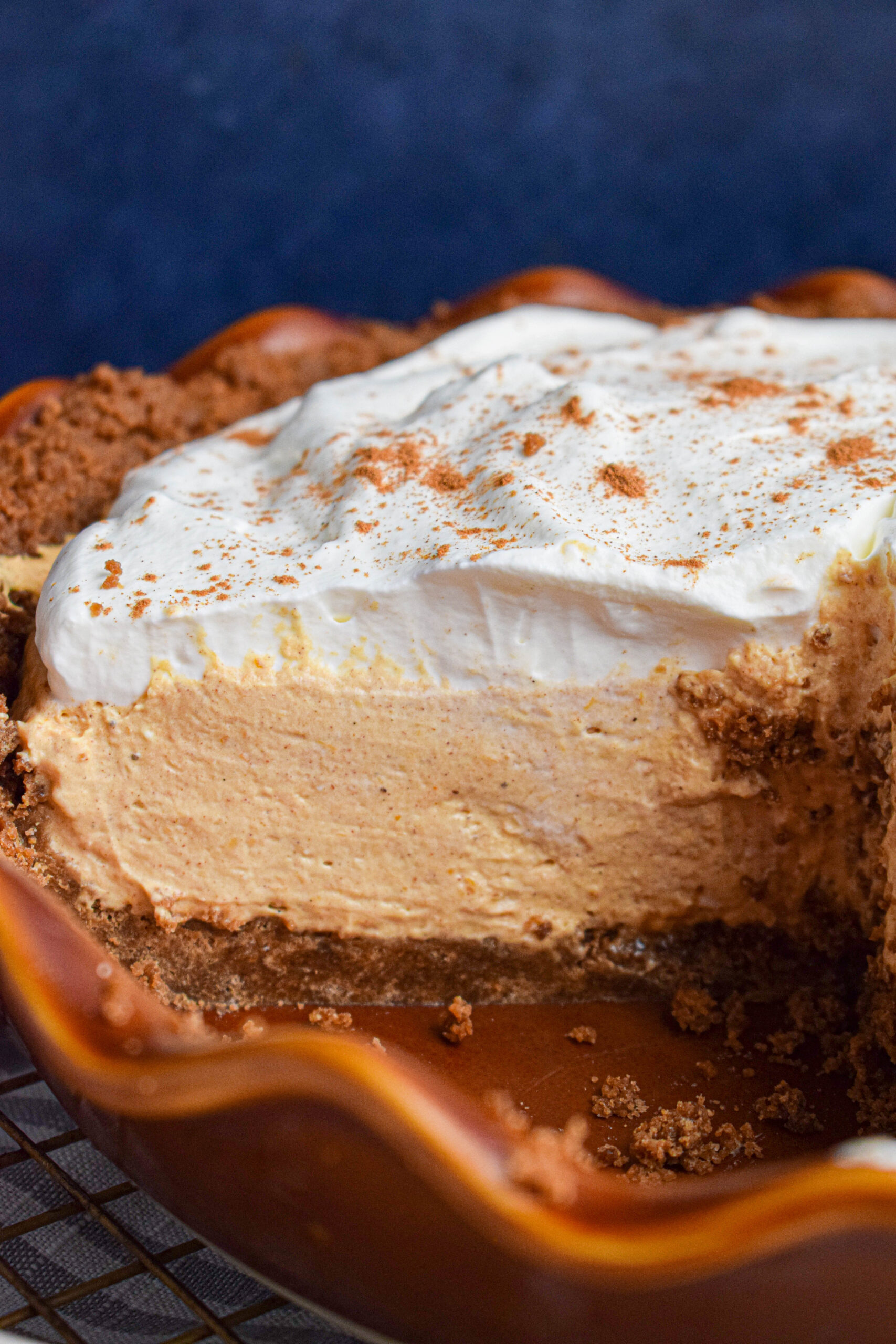 Inside of a sliced no bake pumpkin cheesecake pie showing layers of gingersnap crust, a mousse filling made with no cool whip and topped with whipped cream and cinnamon in a brown pie dish.