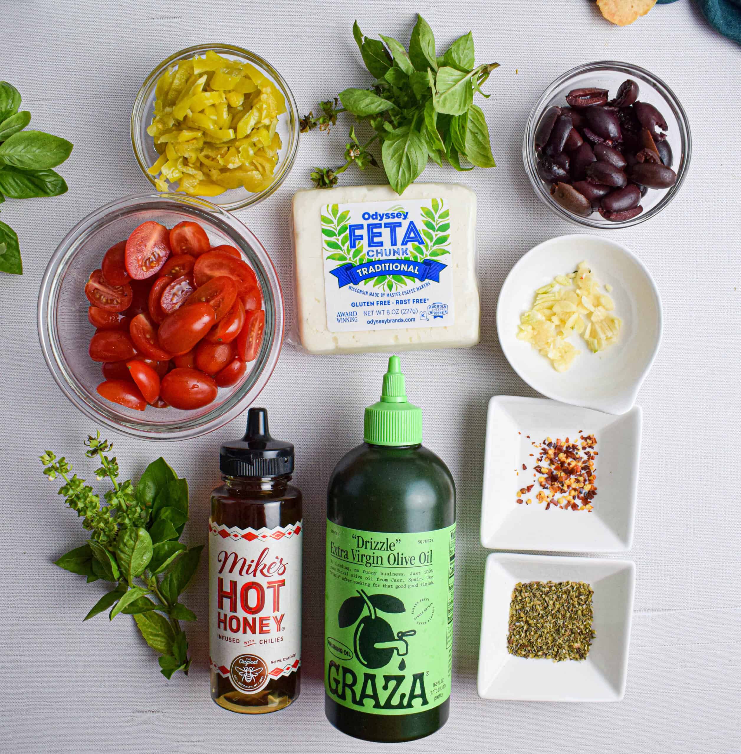 Ingredients for making baked feta dip laid out including, block of feta, olive oil, hot honey, cherry tomatoes, red pepper flakes, garlic, olive, pepperoncini, Italian seasoning and basil. 