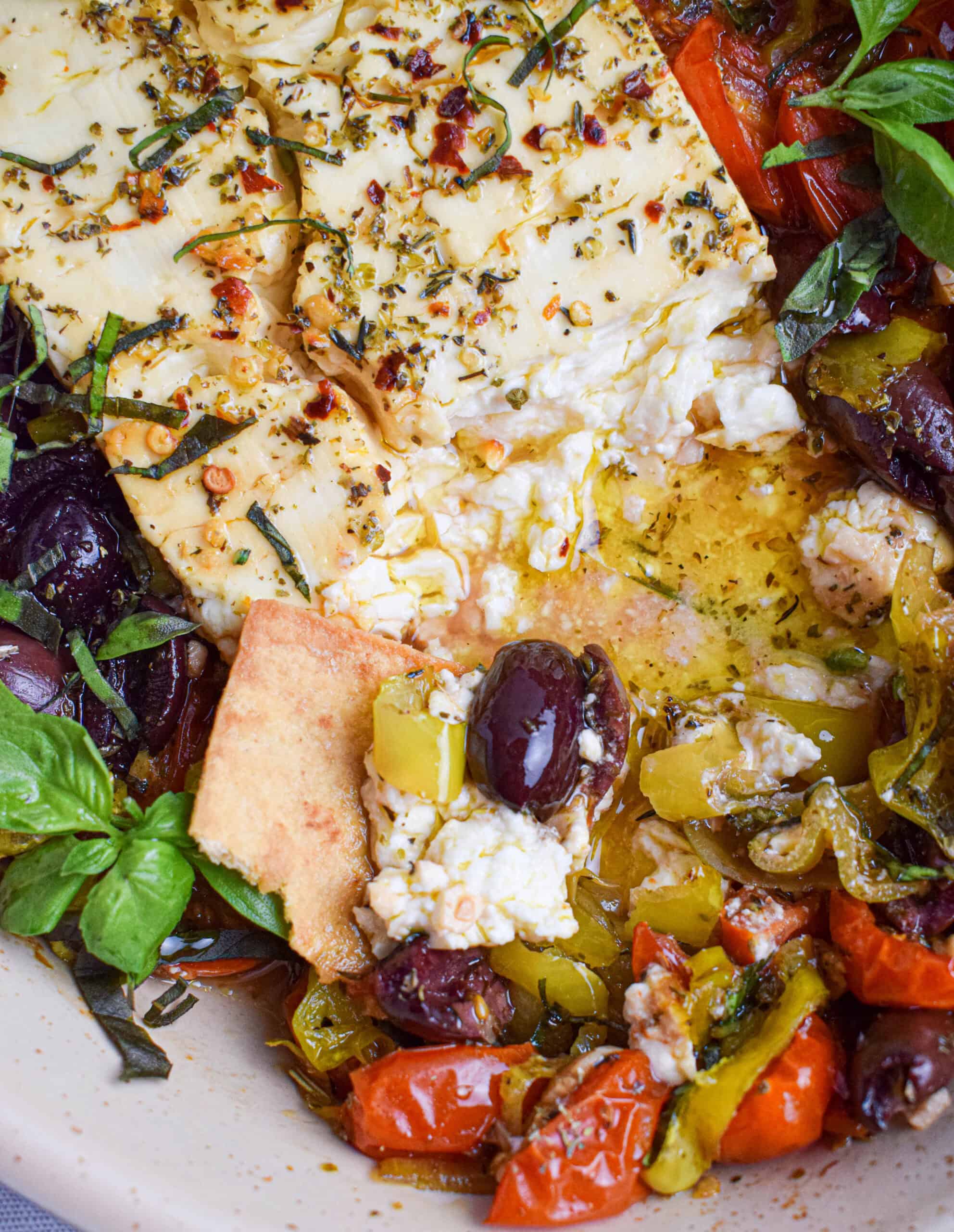 Close up of a pita chip taking in a serving dish filled with spicy baked feta with tomatoes, olives, pepperoncini and basil. 