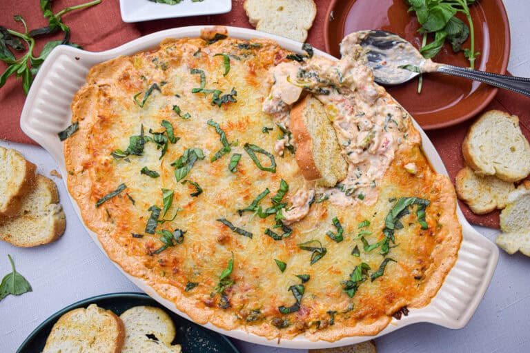 A baked spinach and cheese dip in a casserole dish, topped with melted cheese and fresh basil. A spoon scoops out a portion, revealing the creamy inside. Surrounding the dish are sliced baguettes and a few sprigs of basil on a white tablecloth.