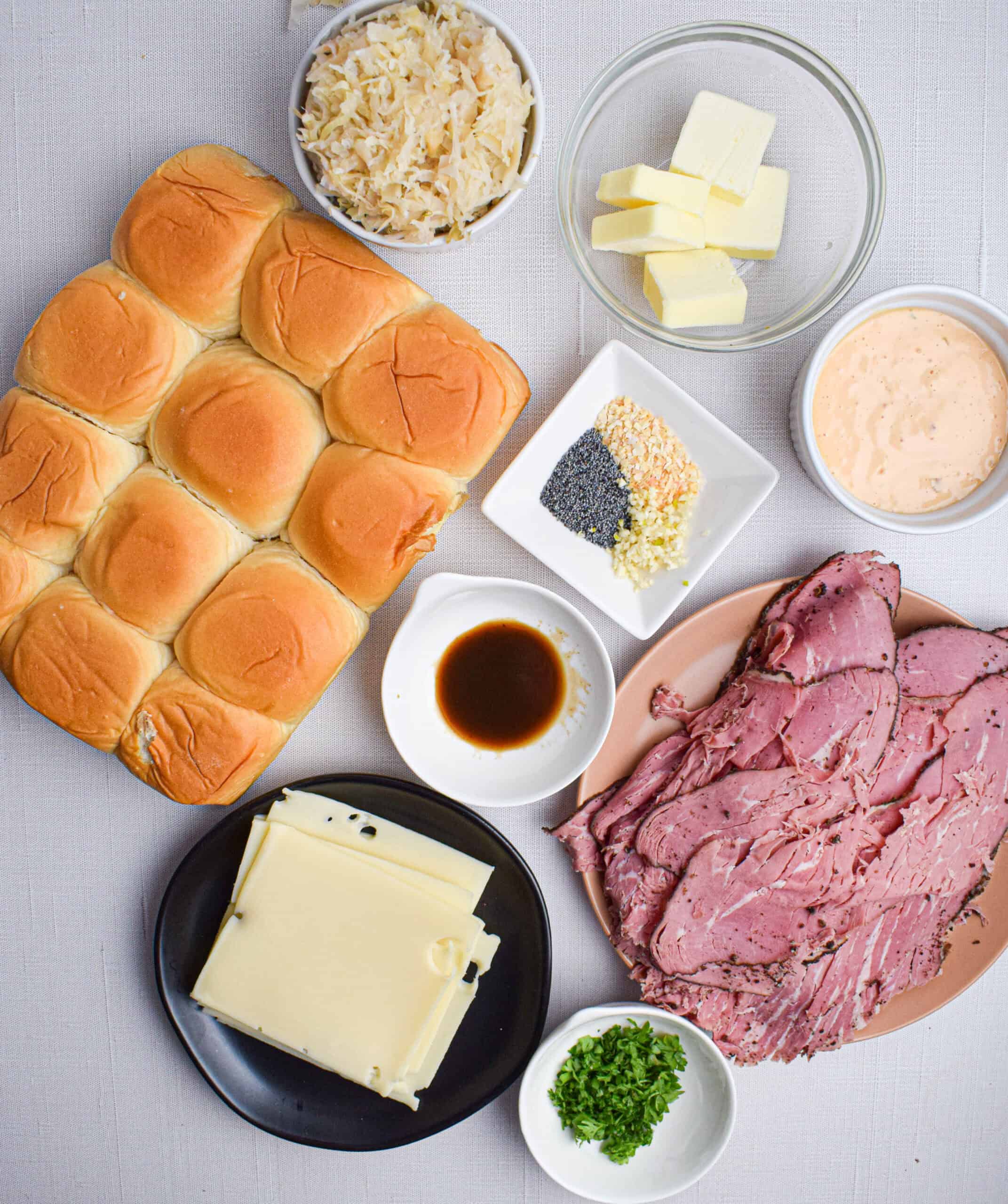 Overhead shot of the ingredients needed to make Reuben sliders laid out: Kings Hawaiian rolls, sliced Swiss cheese, Worcestershire sauce, parsley, garlic, poppy seeds, onion flakes, thousand island dressing, butter and sauerkraut. 