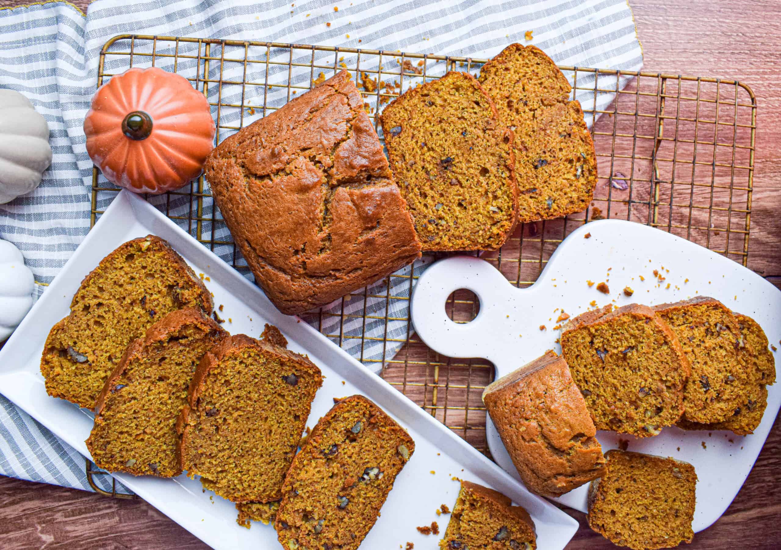 Overhead shot of several loaves of pumpkin bread with pecans on a wire wrack, some slices are on white serving plates and there are some ceramic pumpkins. 