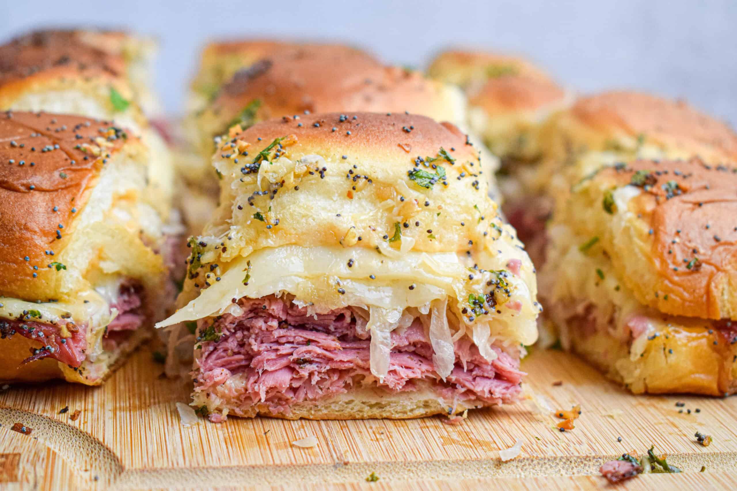 Side shot showing the layers in a Reuben slider of pastrami, sauerkraut and melted cheese on a rolls with a golden topping with poppy seeds and parsley. 