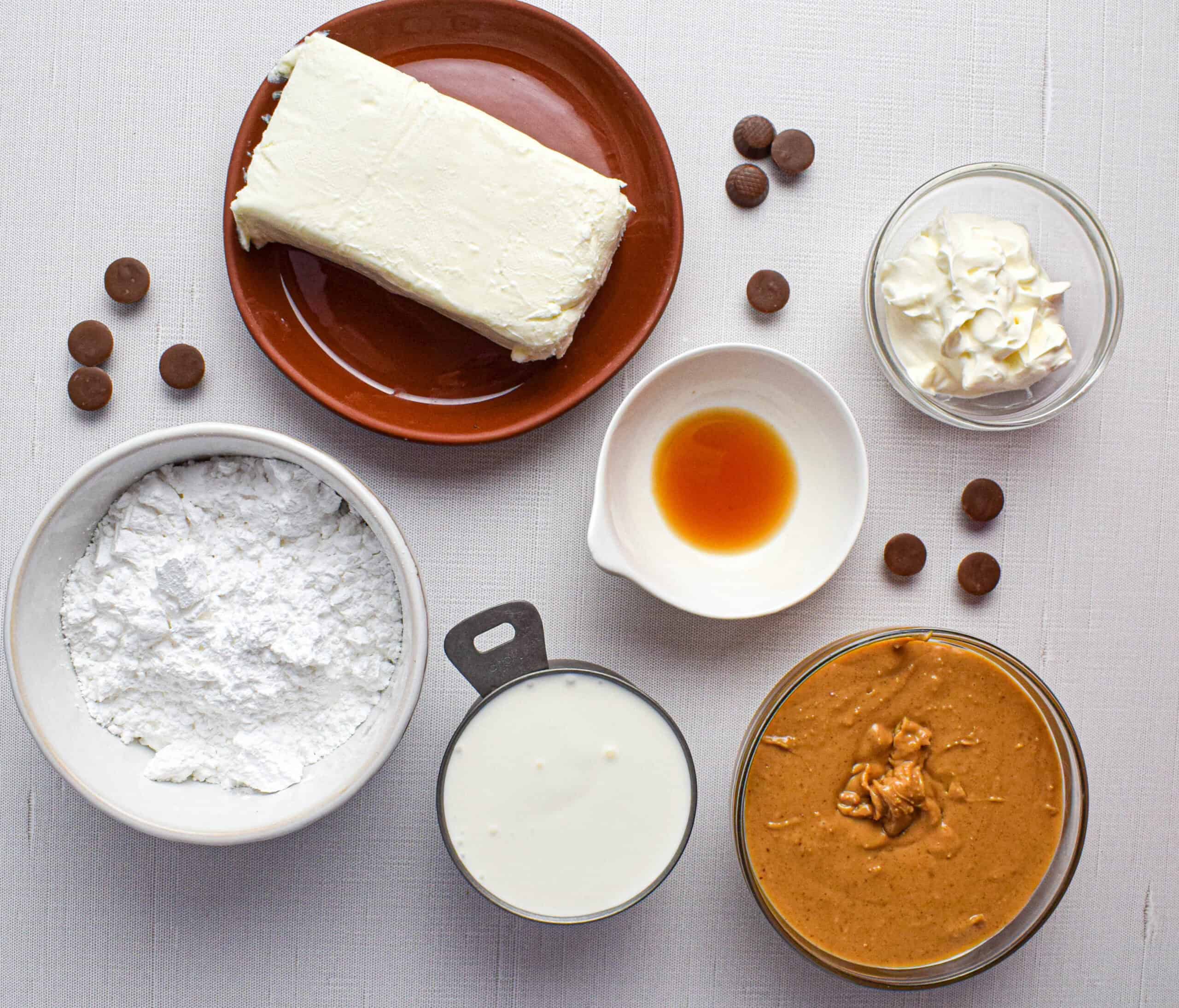 Overhead shot of the ingredients for peanut butter mousse laid out, softened cream cheese, powdered sugar, sour cream, vanilla extract, peanut butter, heavy cream and mini peanut butter cups. 