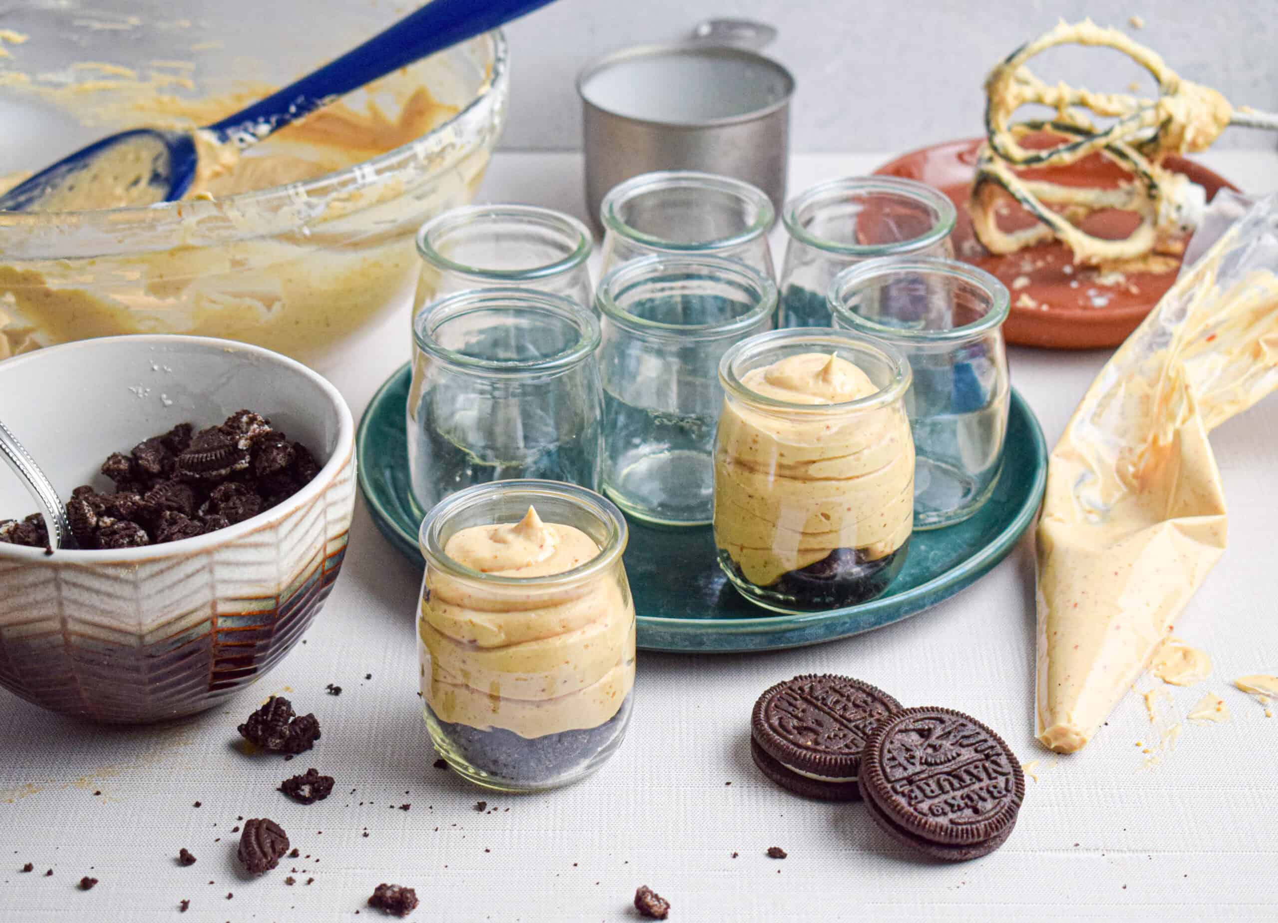 Several small glass jars sit on a green plate with 2 filled with peanut butter mousse with cookie crumbles sitting next to a large mixing bowl filled with the mousse and various baking utensils surround. 