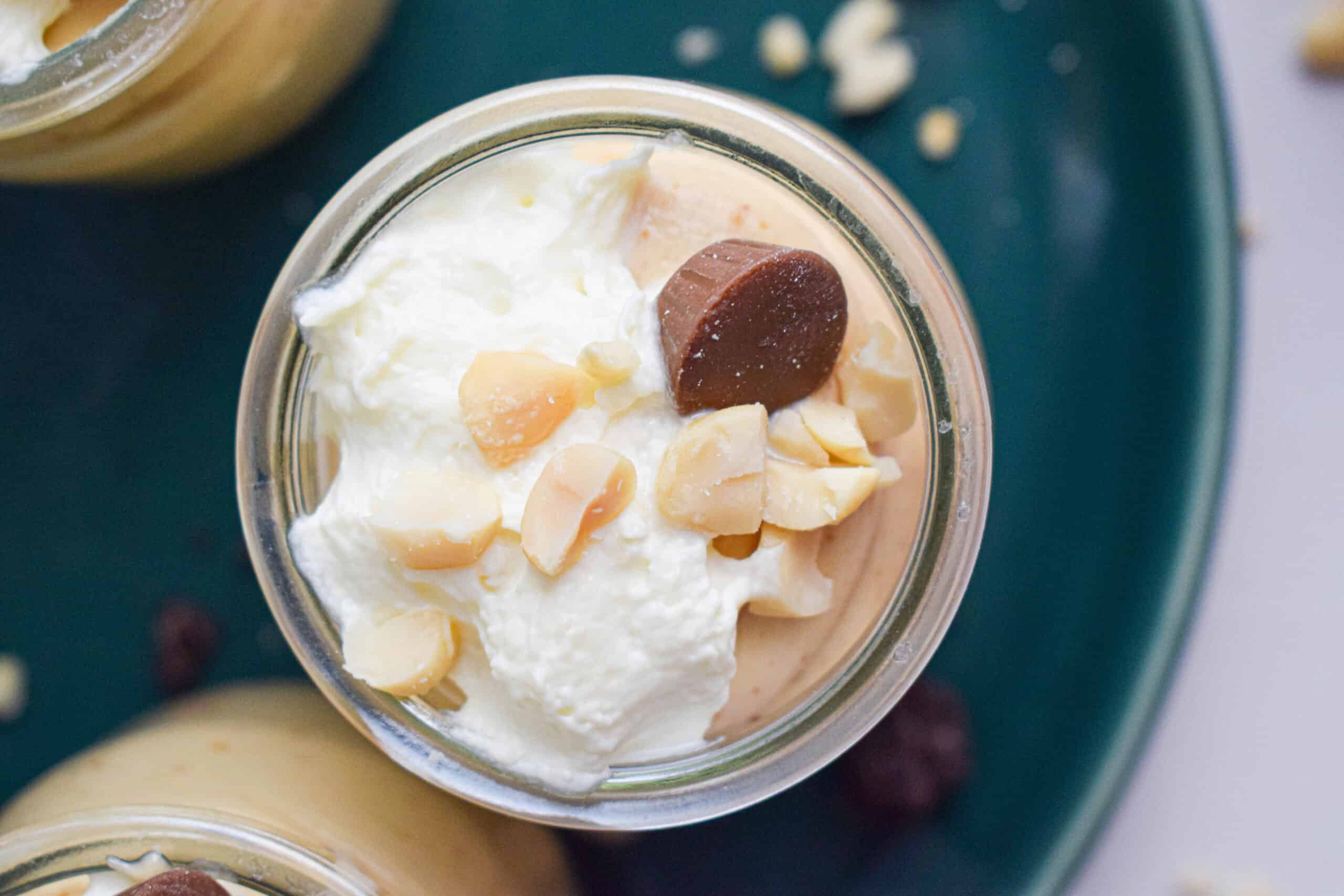 Overhead shot of the top of a jar of peanut mousse with cream, peanuts and a peanut butter cup on a dark green plate. 