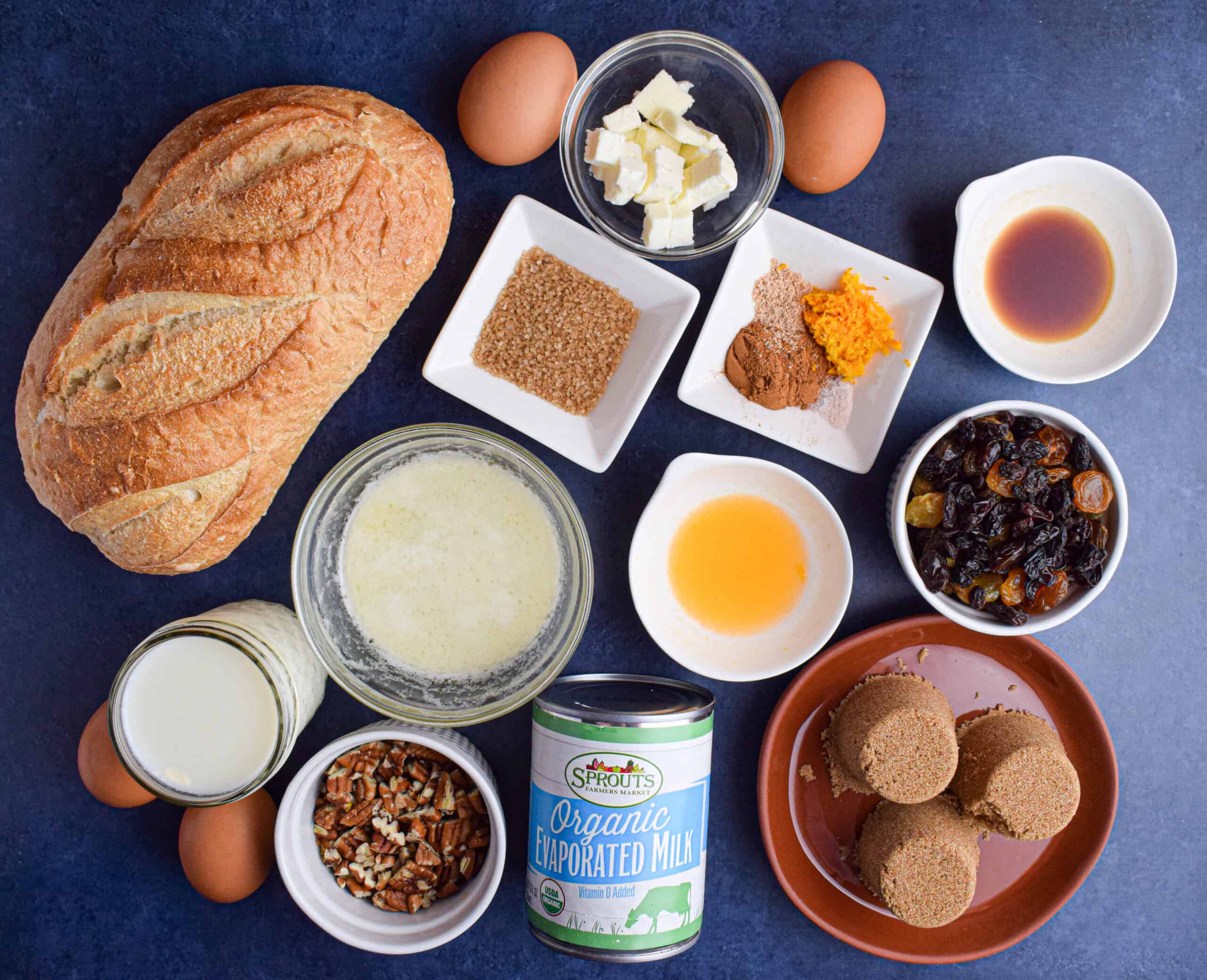 The ingredients for easy sourdough bread pudding laid out including sourdough bread, sugar, butter, eggs, orange zest, raisins, pecans, milk, evaporated milk. 