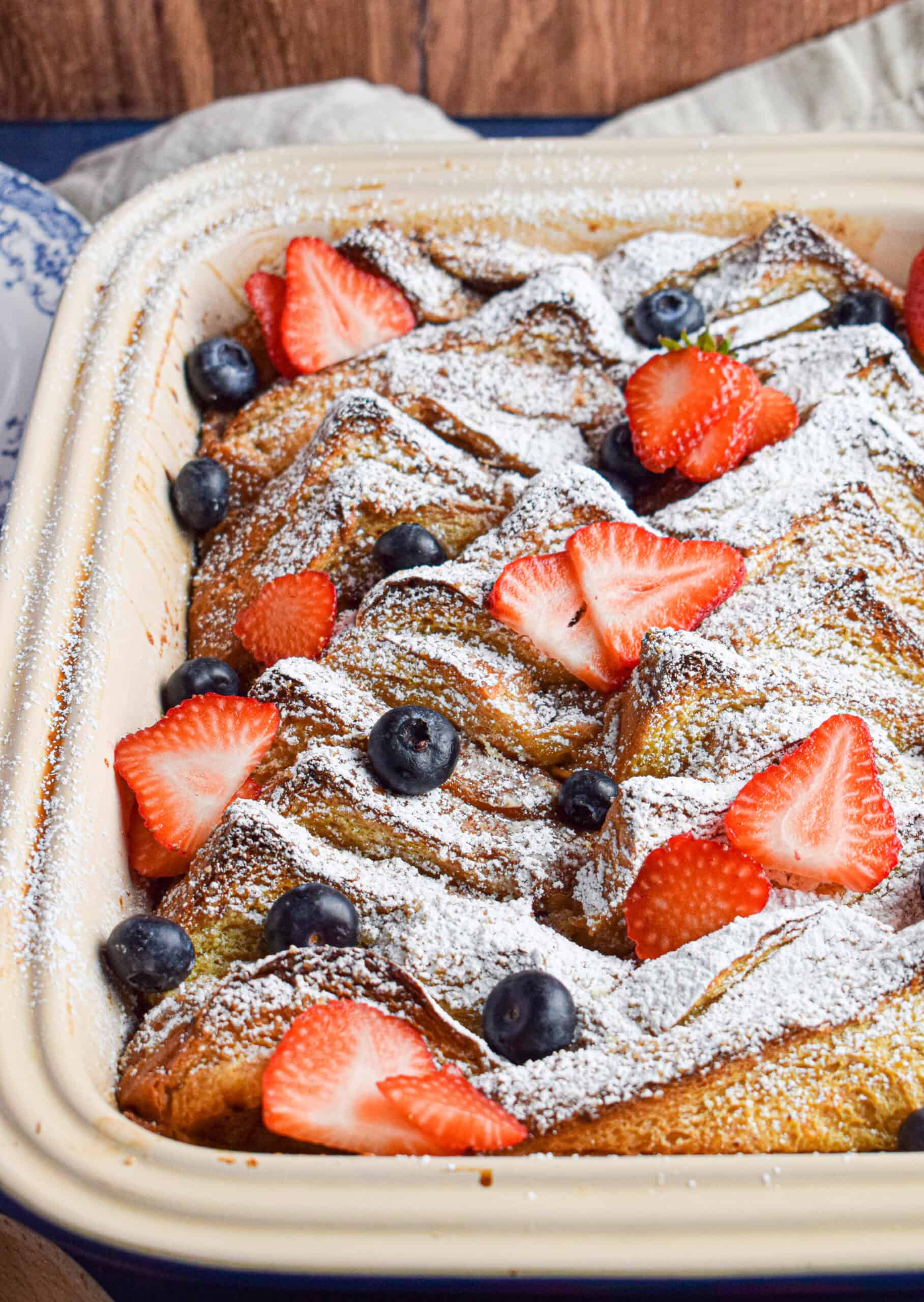 Layered slices in a french toast casserole using brioche bread are baked in a dish topped with powdered sugar, blueberries and strawberries 