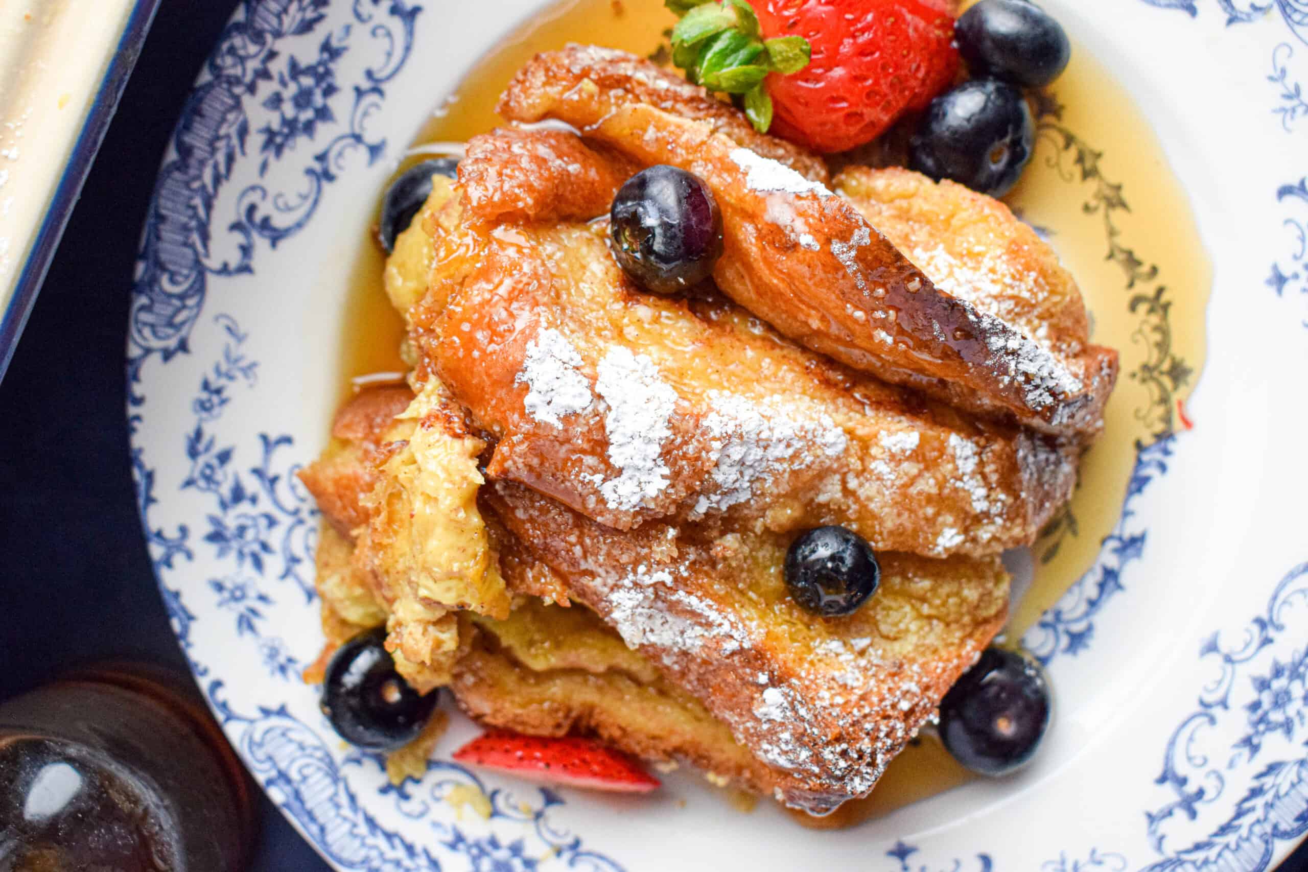 Overhead shot of a serving of layered french toast casserole with brioche that has blueberries, strawberries, maple syrup and powdered sugar. 