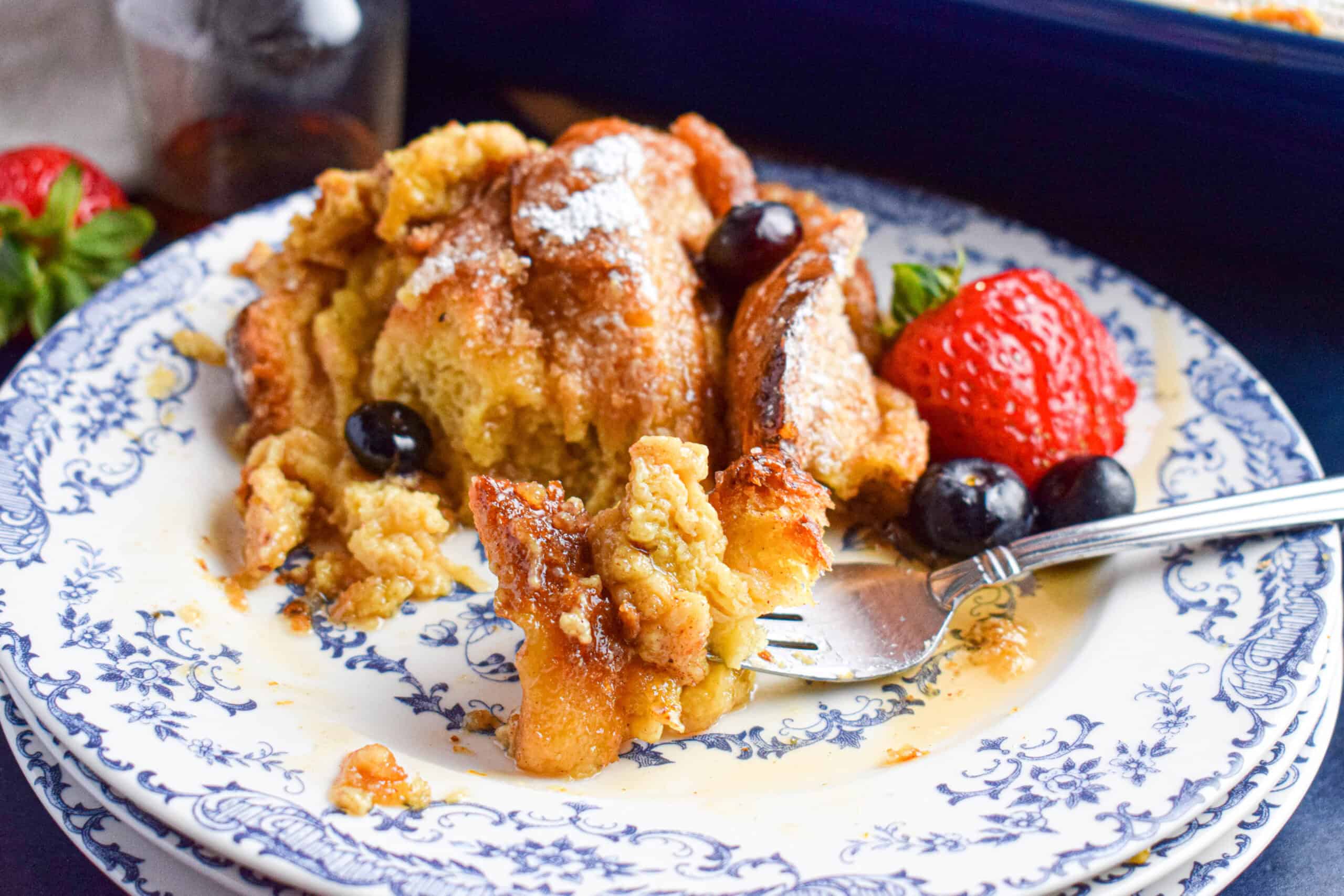 A serving of breakfast bread casserole is on a stack of blue and white plates with berries and a fork taking out a bite. 