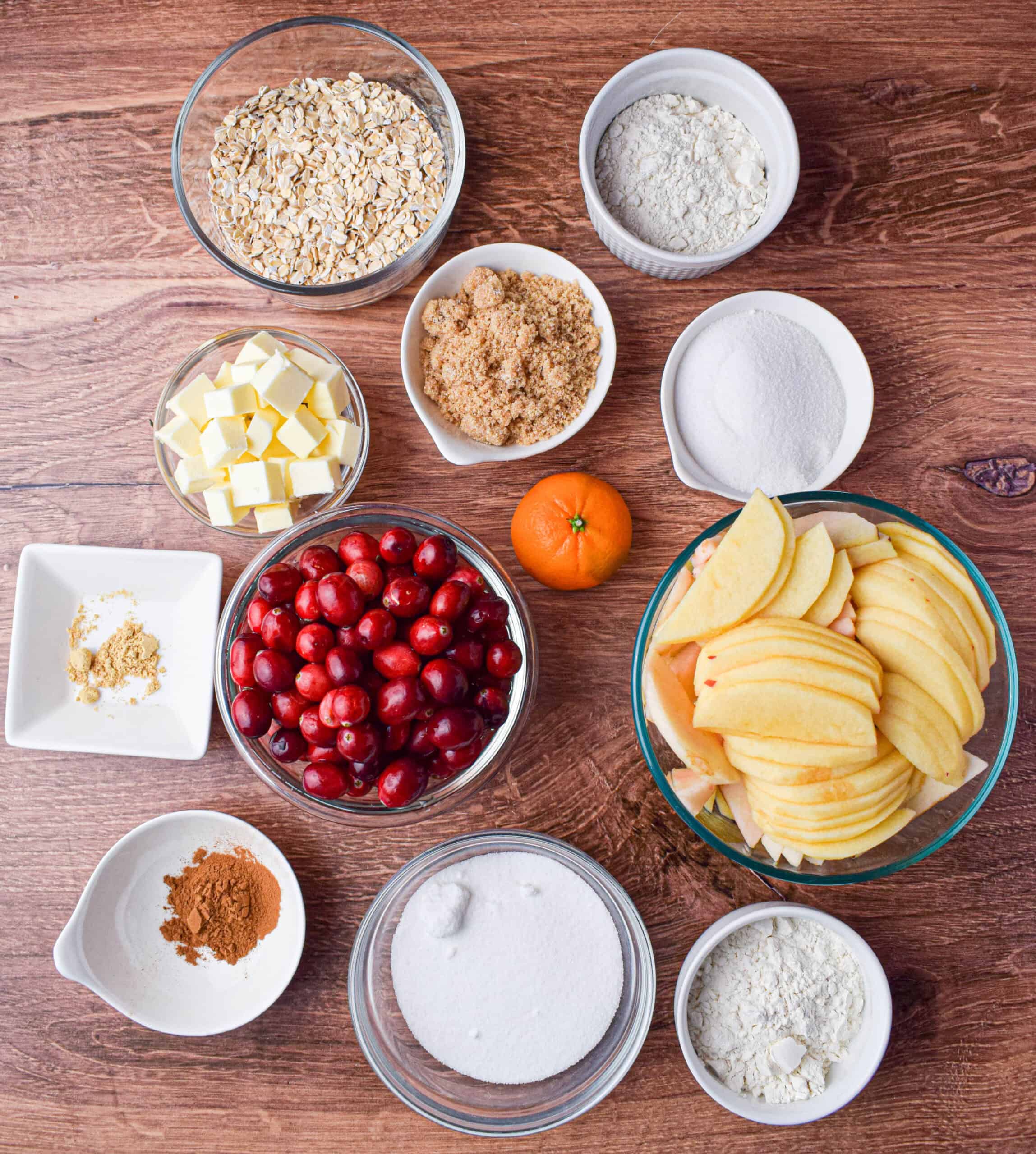 Ingredients laid out for cranberry apple crumble including rolled oats, flour, sugar, orange, cranberries, apples, spices and butter.