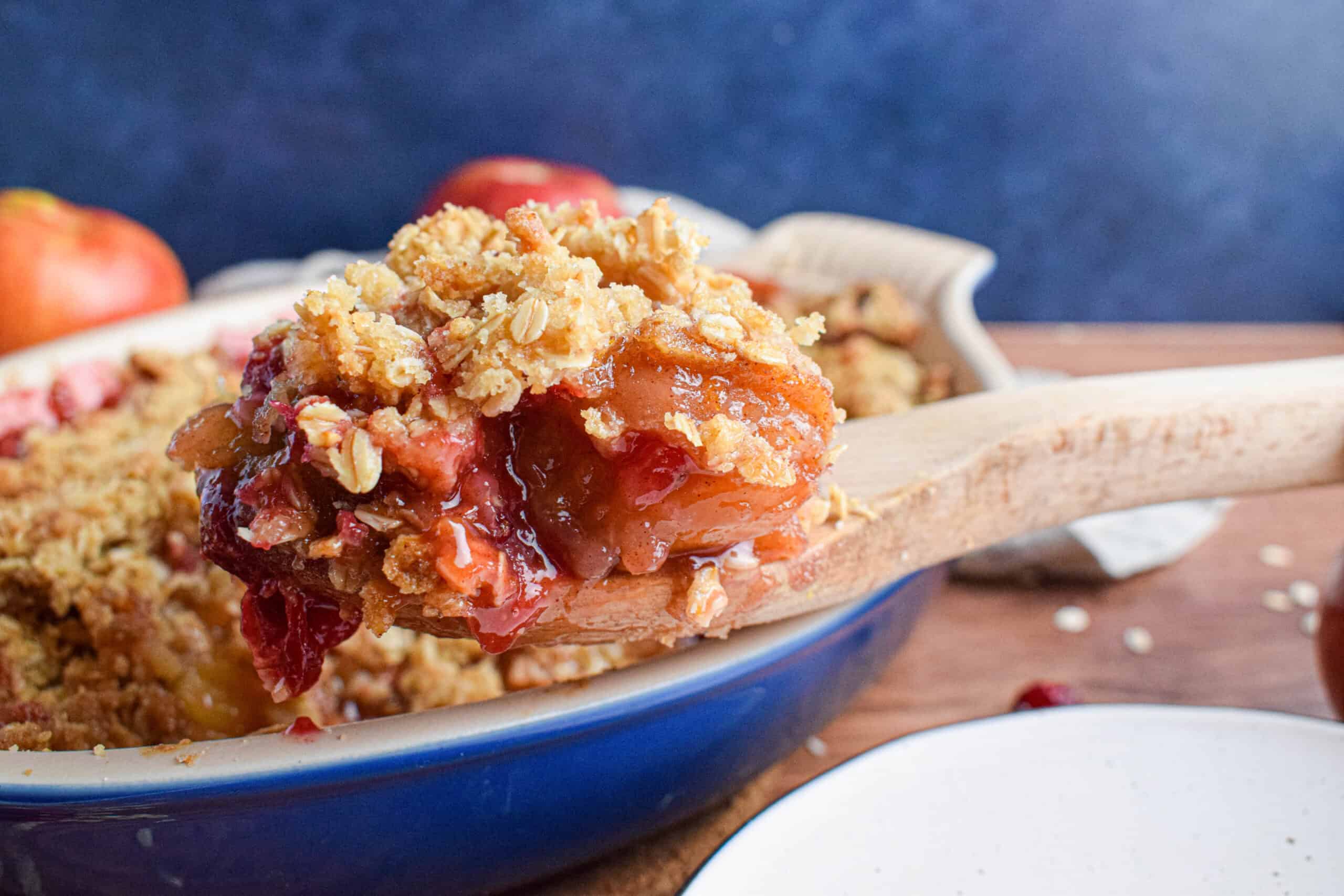 Close up shot of a wooden spoon with a large serving of apple cranberry crisp in front of a blue background.