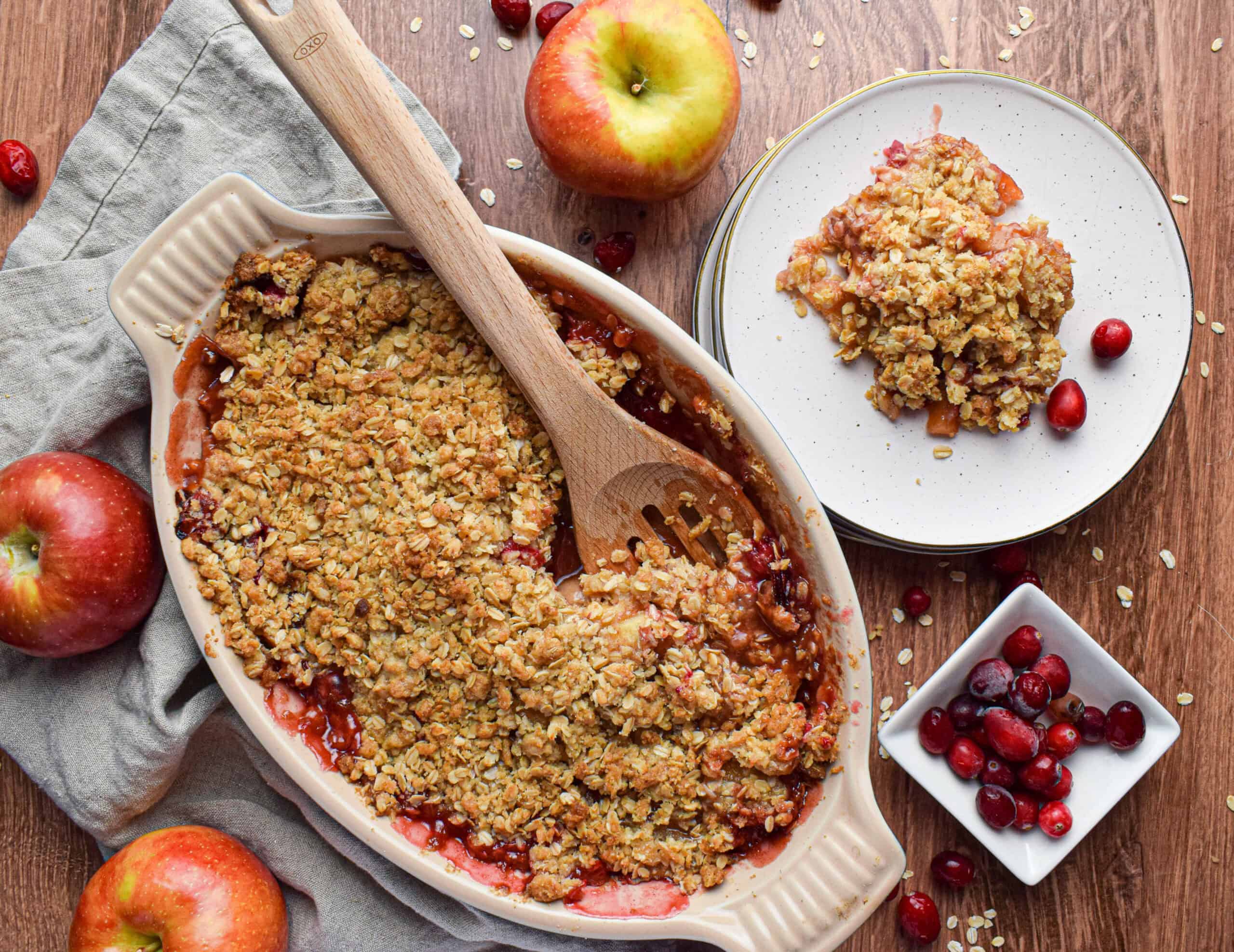 Overhead shot of a serving dish of a crisp apple crumble with a wooden spoon next to a stack of white plates with a serving of crumble, there are also apples and cranberries and a linen.