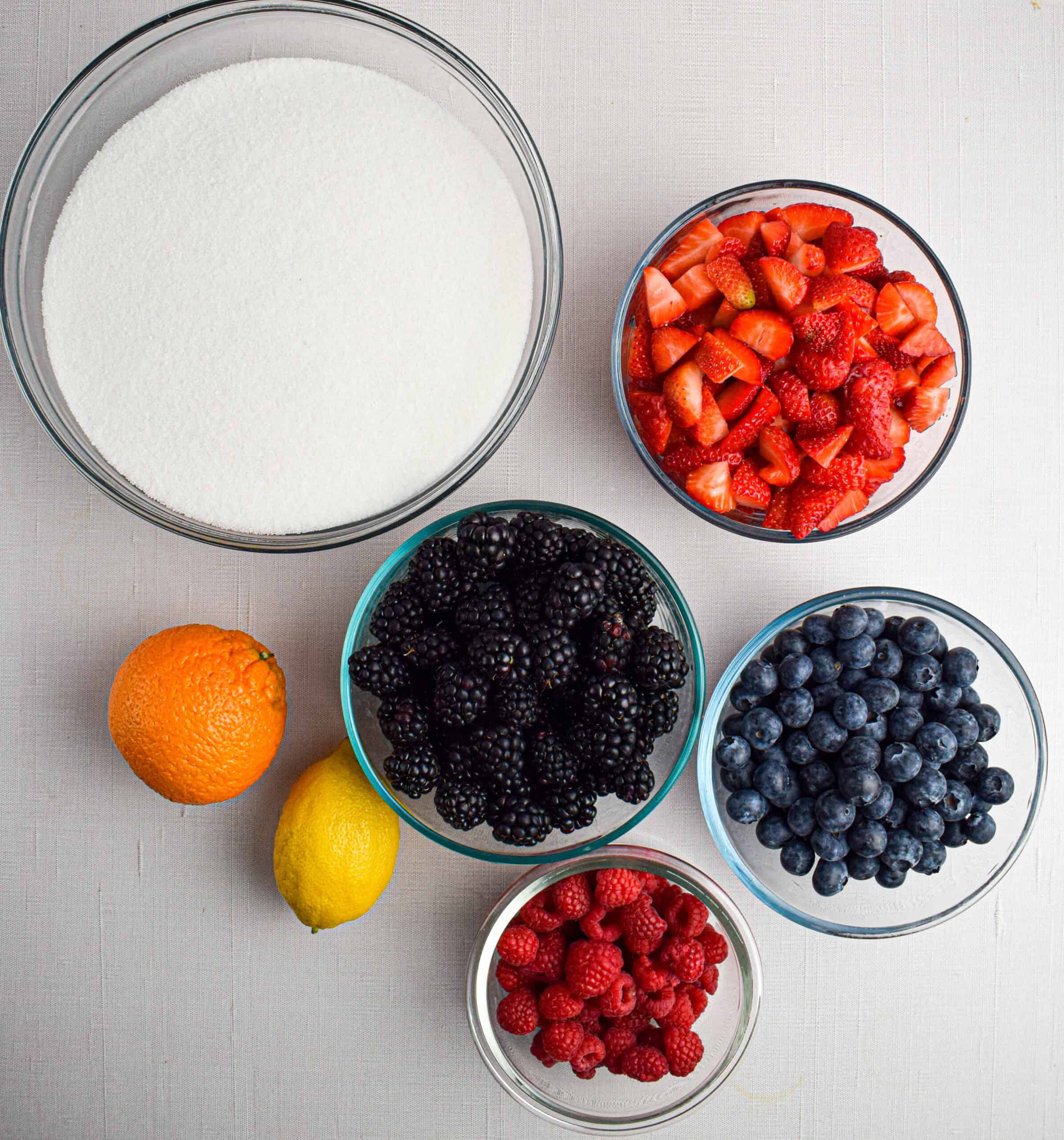 Bowls with the ingredients to make mixed berry jam are laid on a white surface including fresh strawberries, blackberries, blueberries and raspberries along with sugar, a lemon and an orange. 