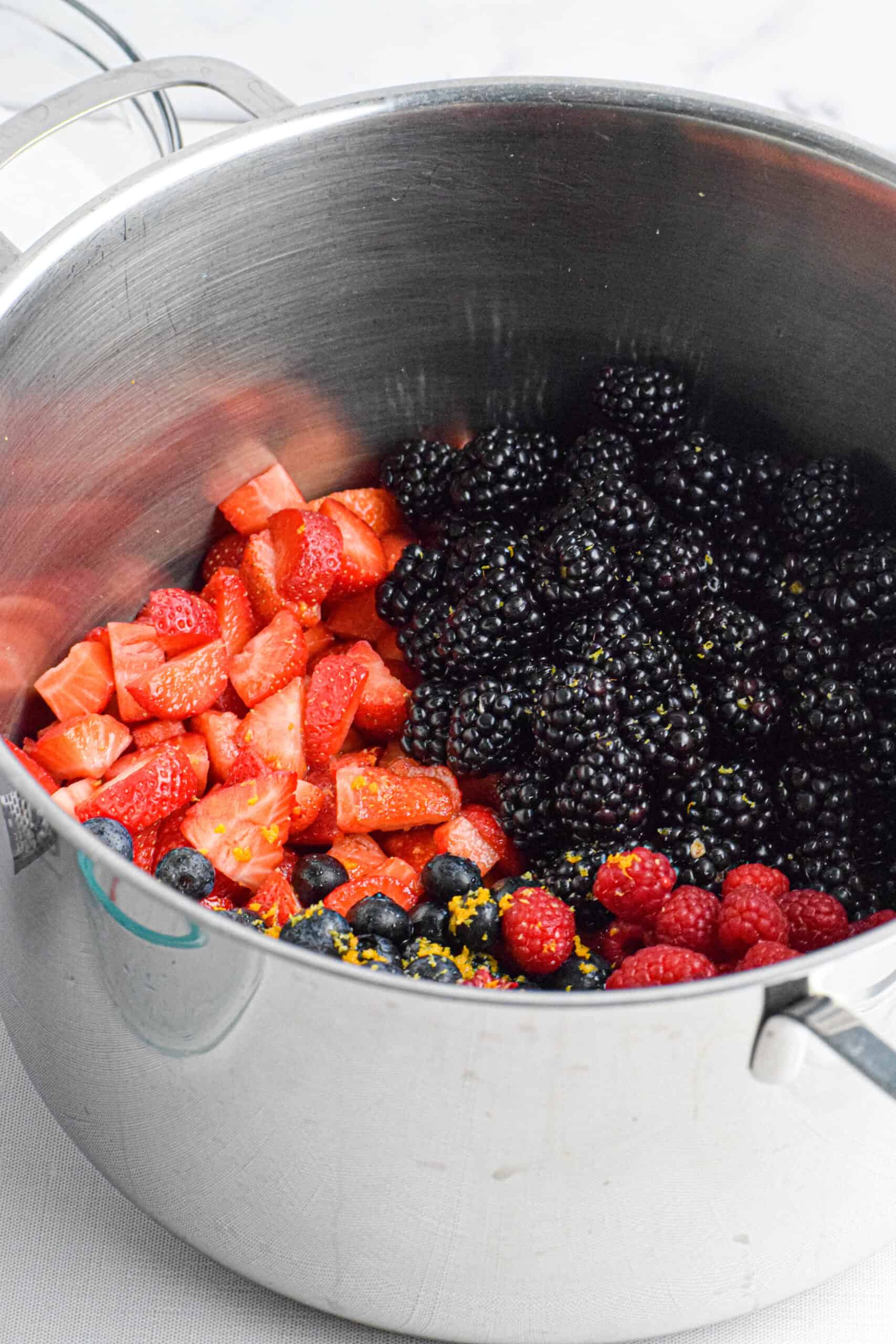 A large stainless steel pot is full of ingredients to make the best homemade berry jam without pectin,  including cut strawberries, blackberries, blueberries, raspberries and citrus zest. 