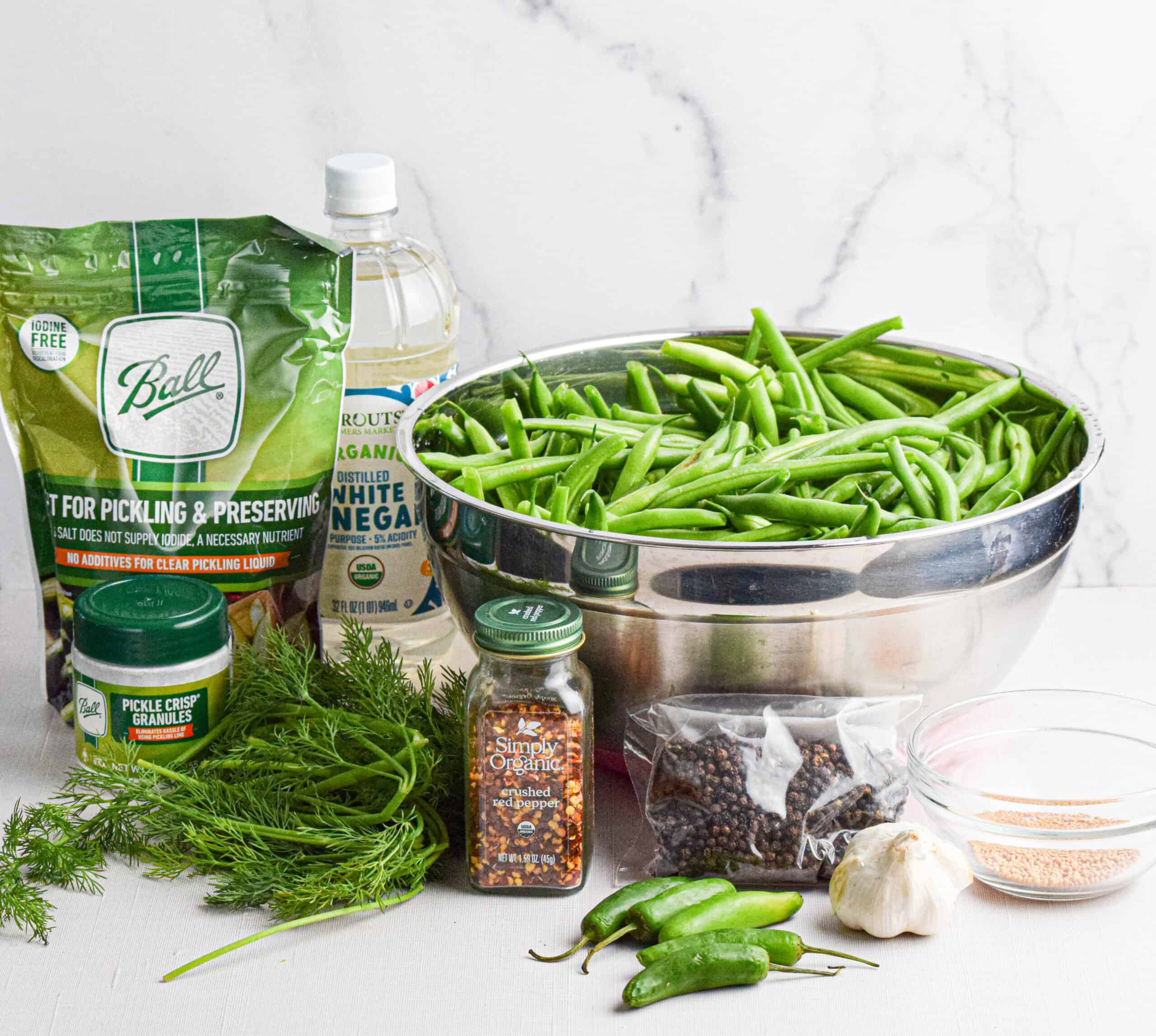 The ingredients needed to pickle green beans are setting on a white backdrop including a large silver bowl of fresh green beans, fresh dill, Serrano peppers, garlic, black peppercorns, mustard seeds, a jar of red pepper flakes, a jar of pickle crips, a bag of pickling salt and a jar of white vinegar. 